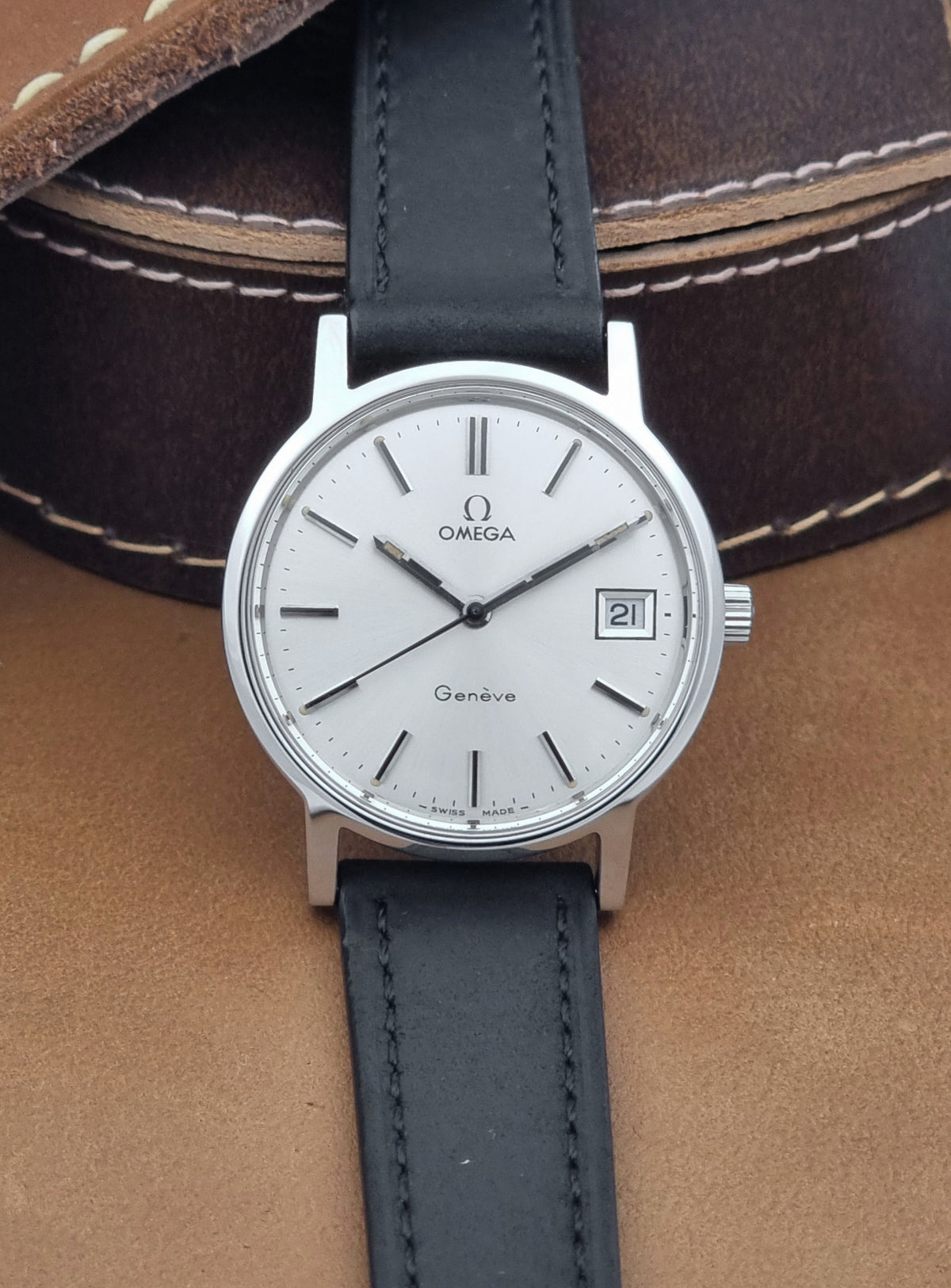 A Vintage Omega Geneve 136.0098 watch, white dial, and black leather strap, resting on a brown leather surface,