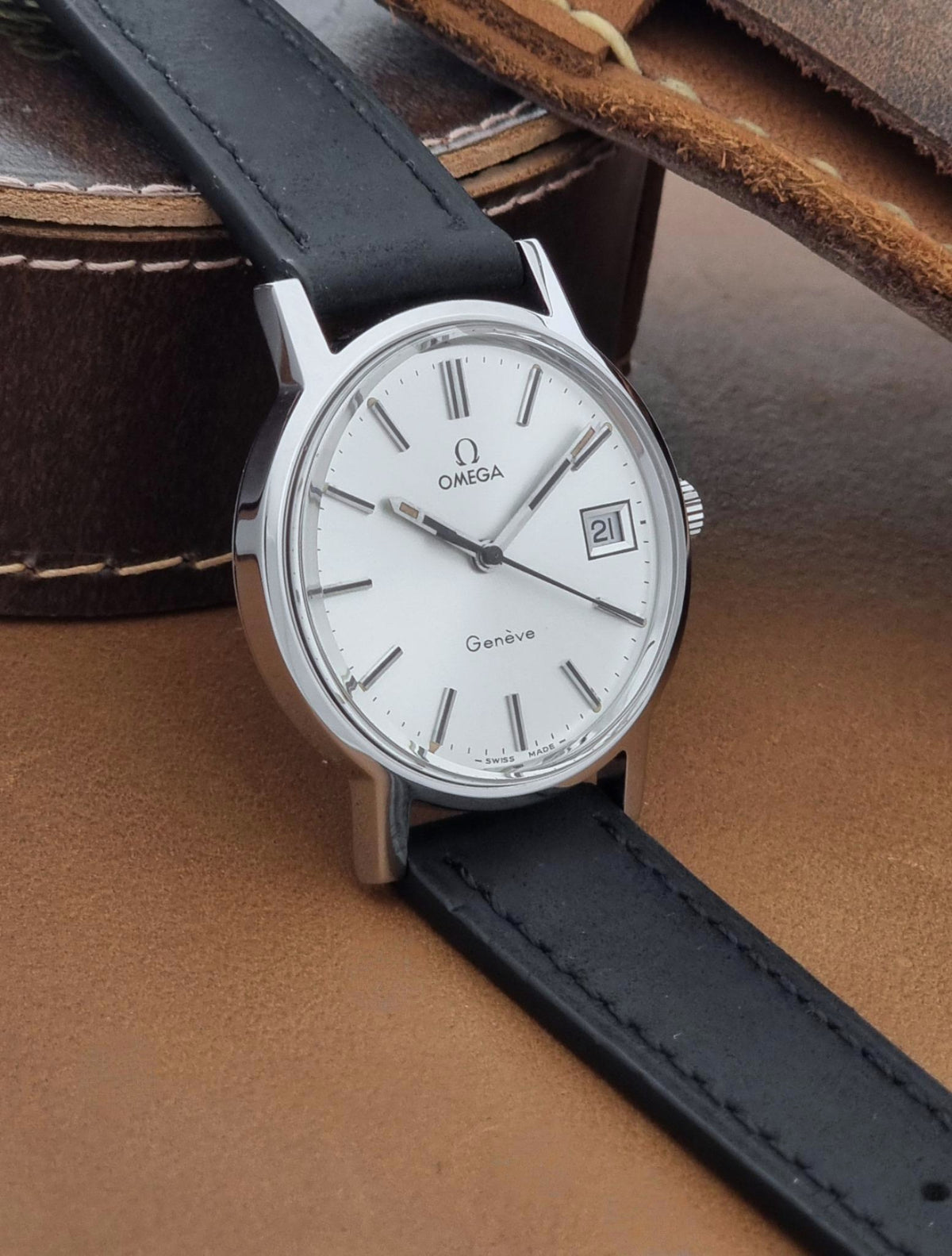 A Vintage Omega Geneve 136.0098 watch, white dial, and black leather strap, resting on a brown leather surface.