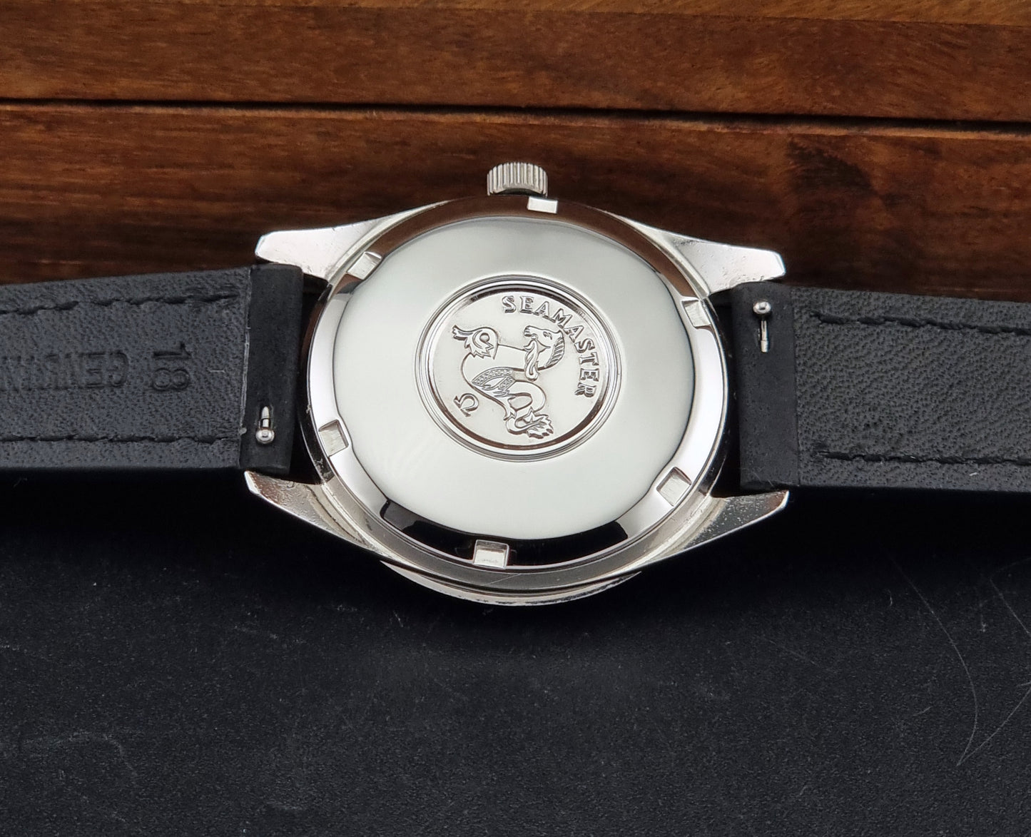 Close-up of the back of a Seamaster wristwatch with a silver-toned metal case,  and a black leather strap on a dark surface with a wooden background.