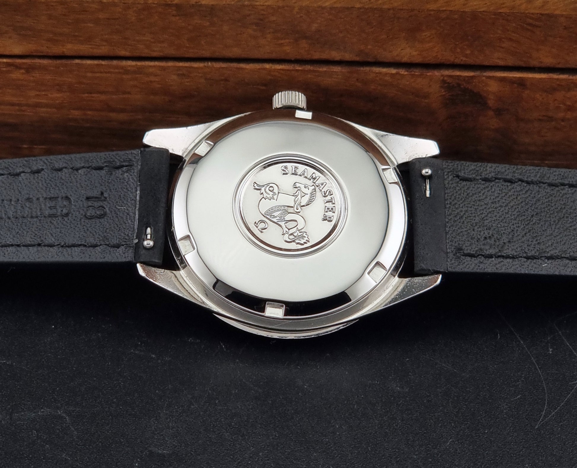 Close-up of the back of a Seamaster wristwatch with a silver-toned metal case,  and a black leather strap on a dark surface with a wooden background.