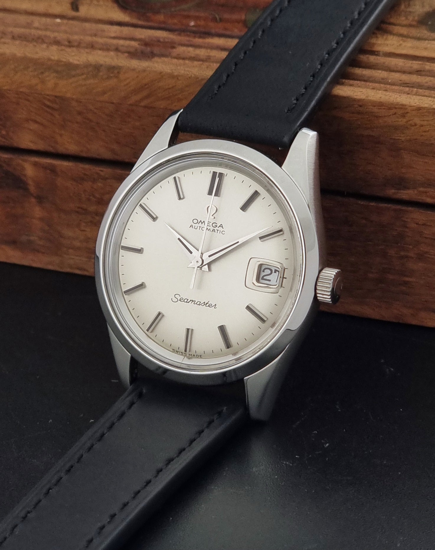 An Omega Seamaster wristwatch with a silver-tone stainless steel case, cream-colored dial, and a black leather strap, resting on a dark surface with a wooden background.