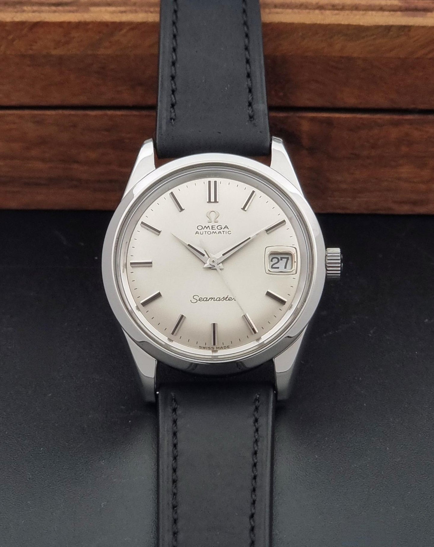 Omega Seamaster wristwatch with a silver-toned stainless steel case, white dial, and black leather strap, displayed on a dark surface with a wooden background.