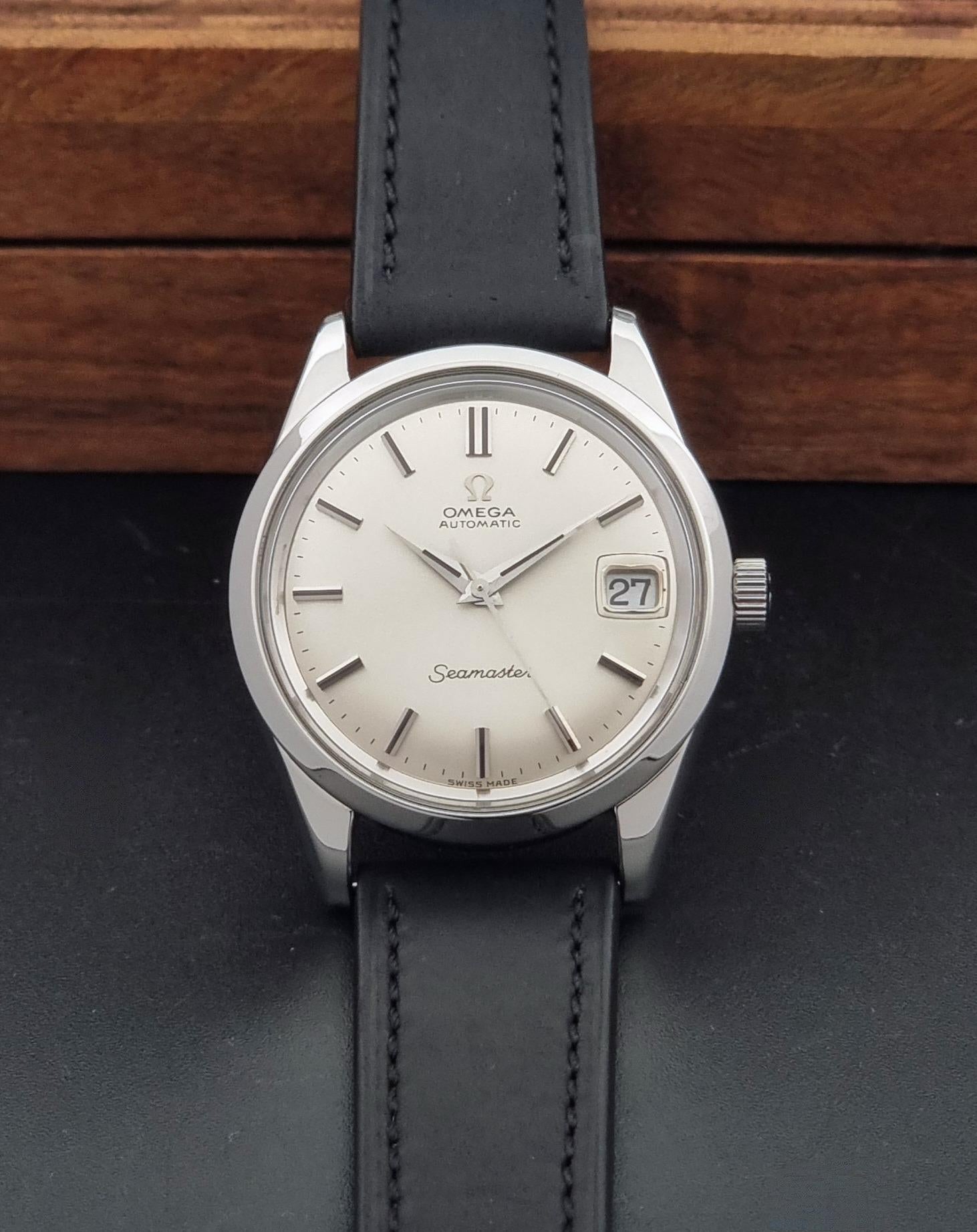 Omega Seamaster wristwatch with a silver-toned stainless steel case, white dial, and black leather strap, displayed on a dark surface with a wooden background.