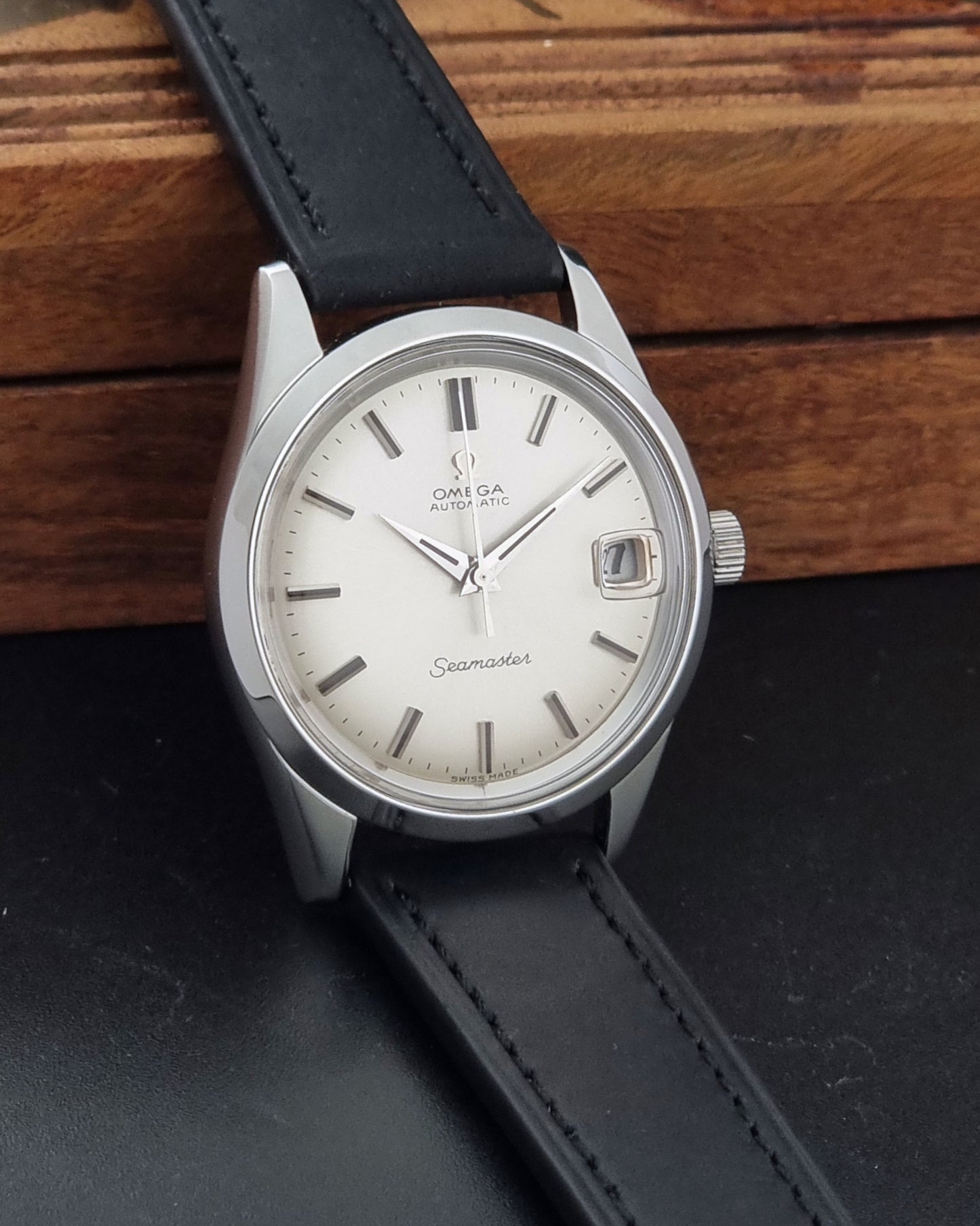 An Omega Seamaster wristwatch with a silver-toned stainless steel case, a white dial, and a black leather strap, resting on a dark surface with a wooden background.