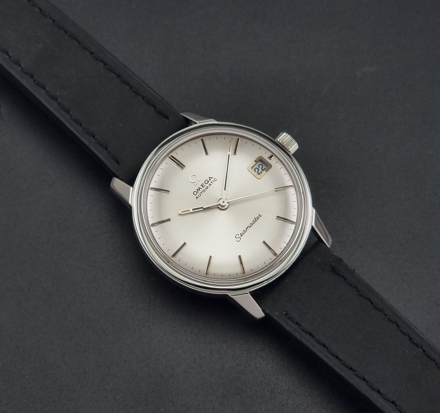 A Vintage Omega Seamaster watch 166.037 wristwatch with a silver-toned stainless steel case, a white dial with a date window, and a black leather strap, shown on a dark gray textured background.