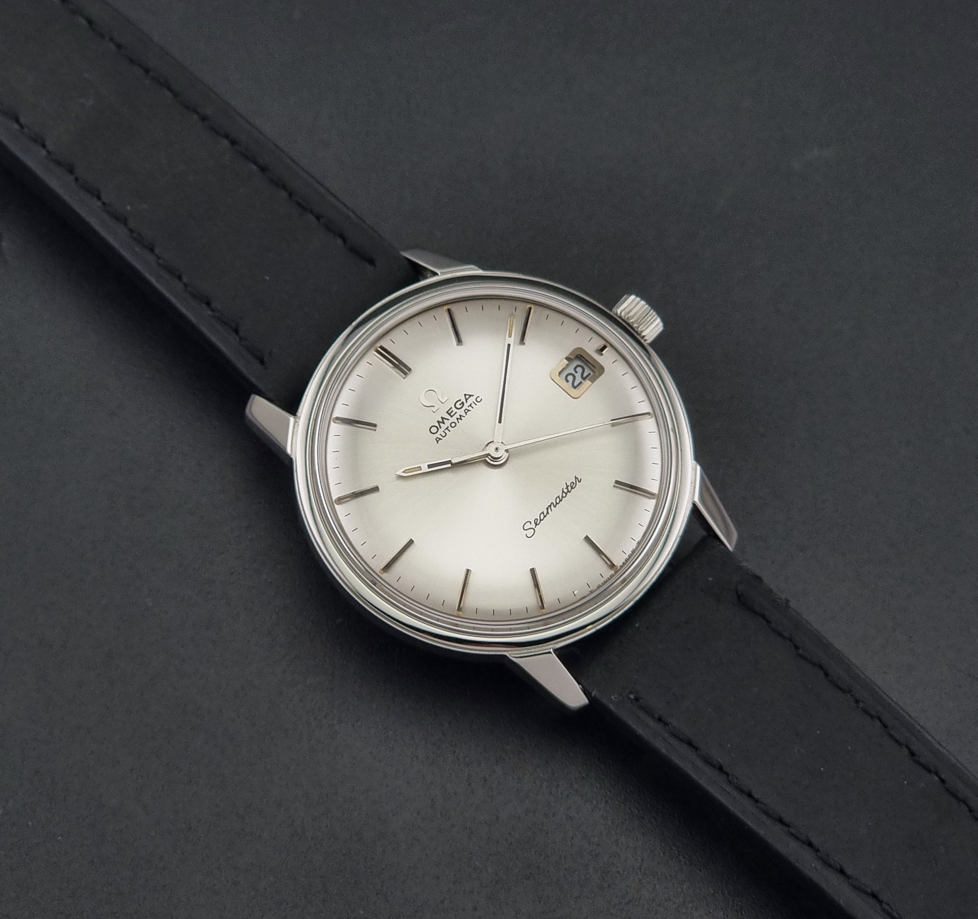 A Vintage Omega Seamaster watch 166.037 wristwatch with a silver-toned stainless steel case, a white dial with a date window, and a black leather strap, shown on a dark gray textured background.