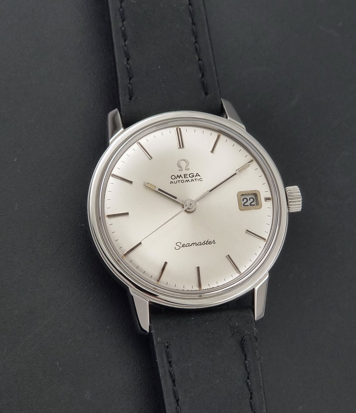 An Omega Seamaster wristwatch with a silver-toned stainless steel case, a white dial, and a black leather strap, shown on a dark textured background.