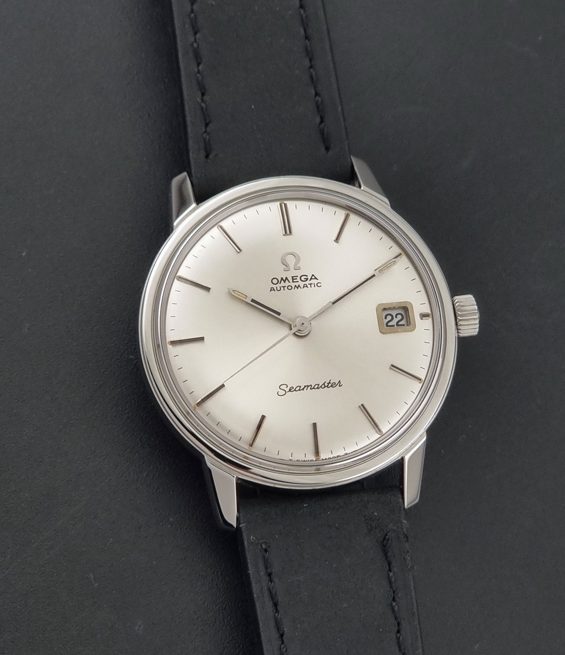An Omega Seamaster wristwatch with a silver-toned stainless steel case, a white dial, and a black leather strap, shown on a dark textured background.