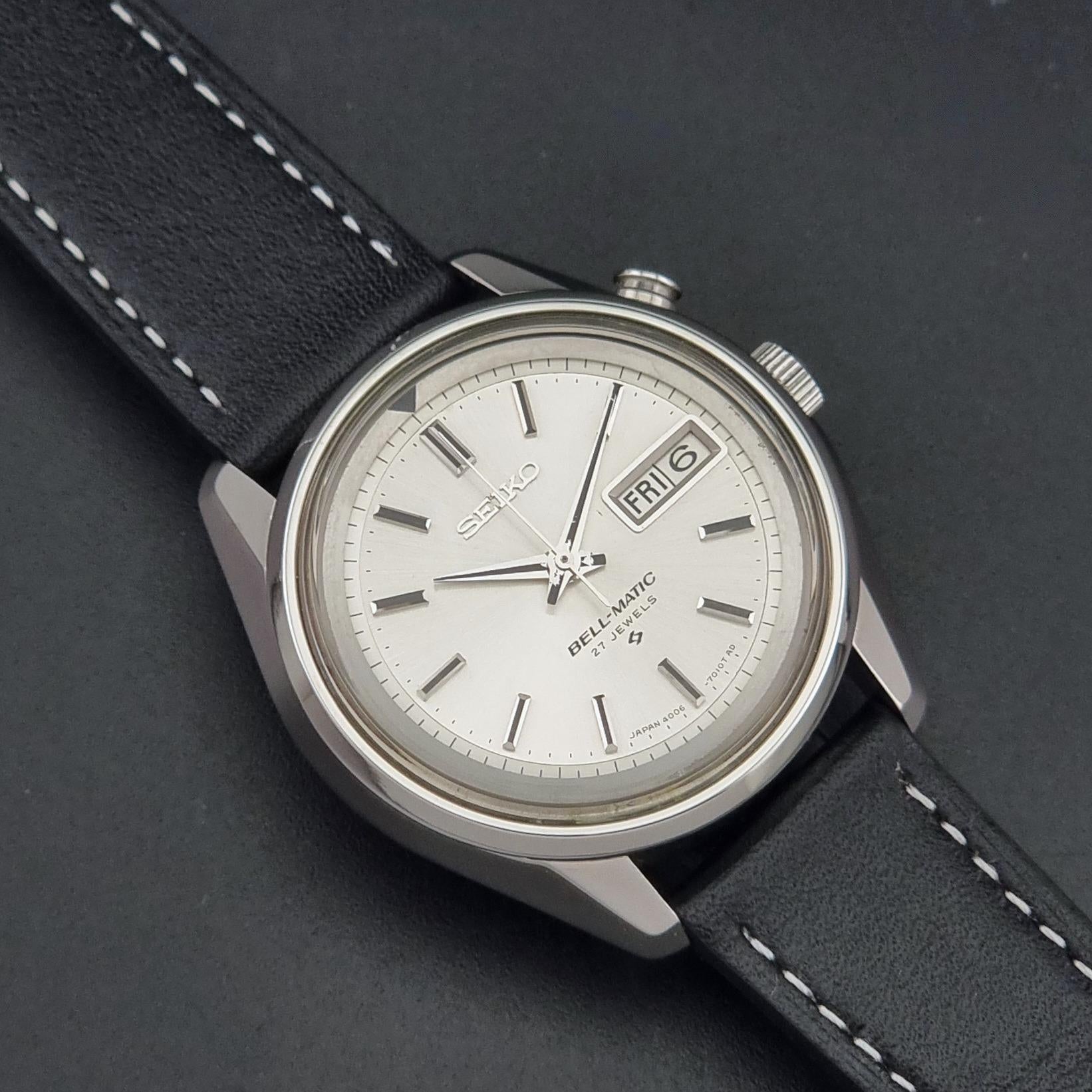 A vintage Seiko-Bell-Matic 4006 wristwatch with a silver-toned case, white dial, and black leather strap with white stitching, shown on a dark textured background.