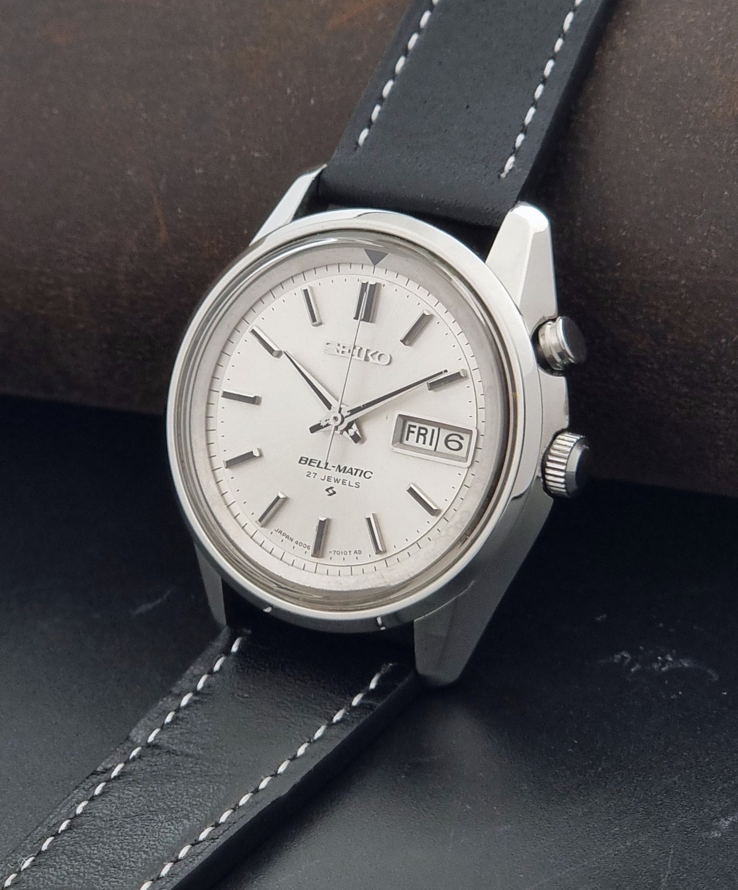 A Vintage Seiko Bell Matic Alarm 4006-7010 automatic wristwatch with a silver-toned metal case, white dial, and black leather strap with white stitching, resting on a dark textured surface.