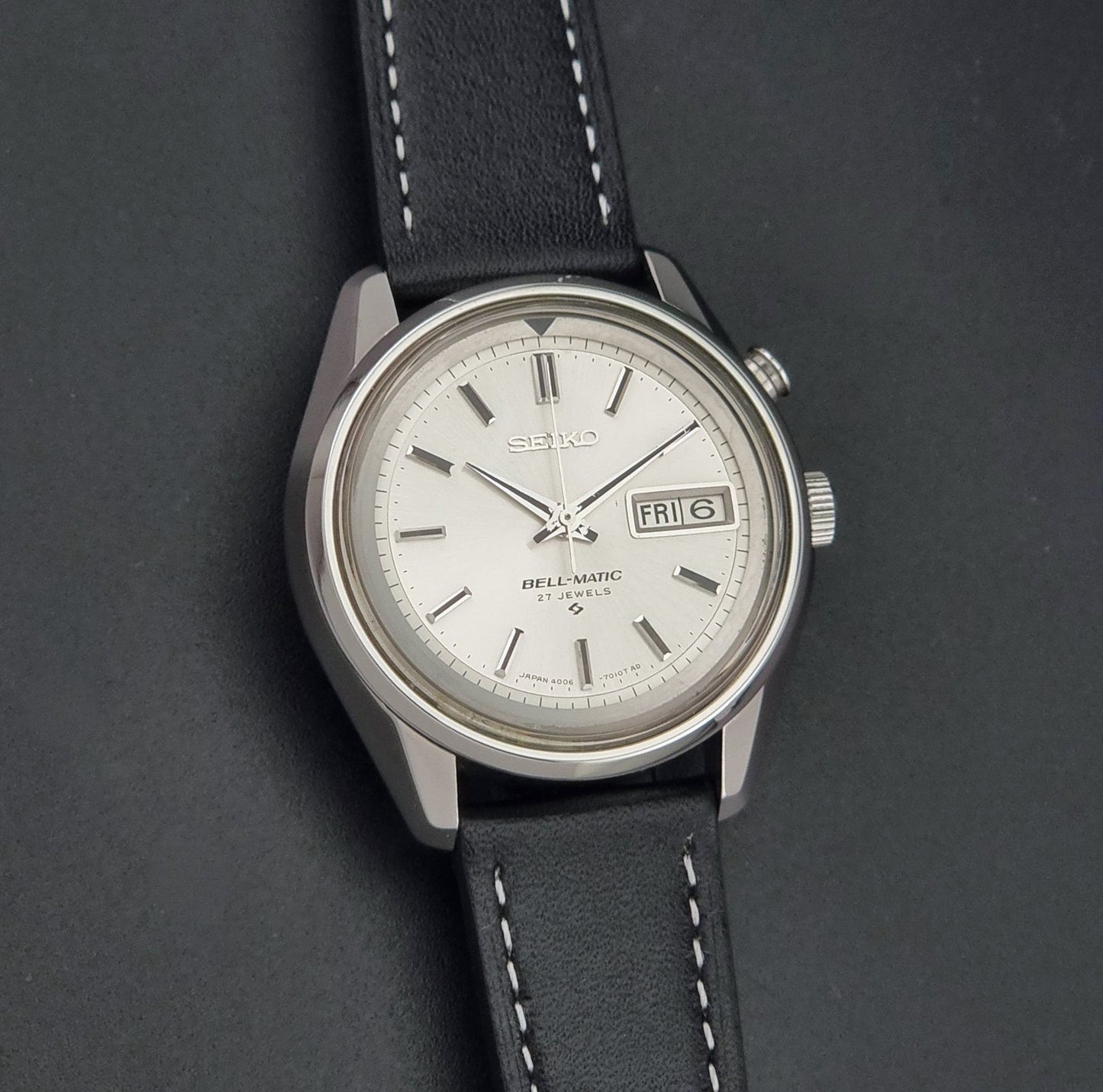 Vintage Seiko Wristwatch Bell Matic Alarm 4006-7010, a white dial with black stick indices, and a black leather strap with white stitching, shown on a dark background.