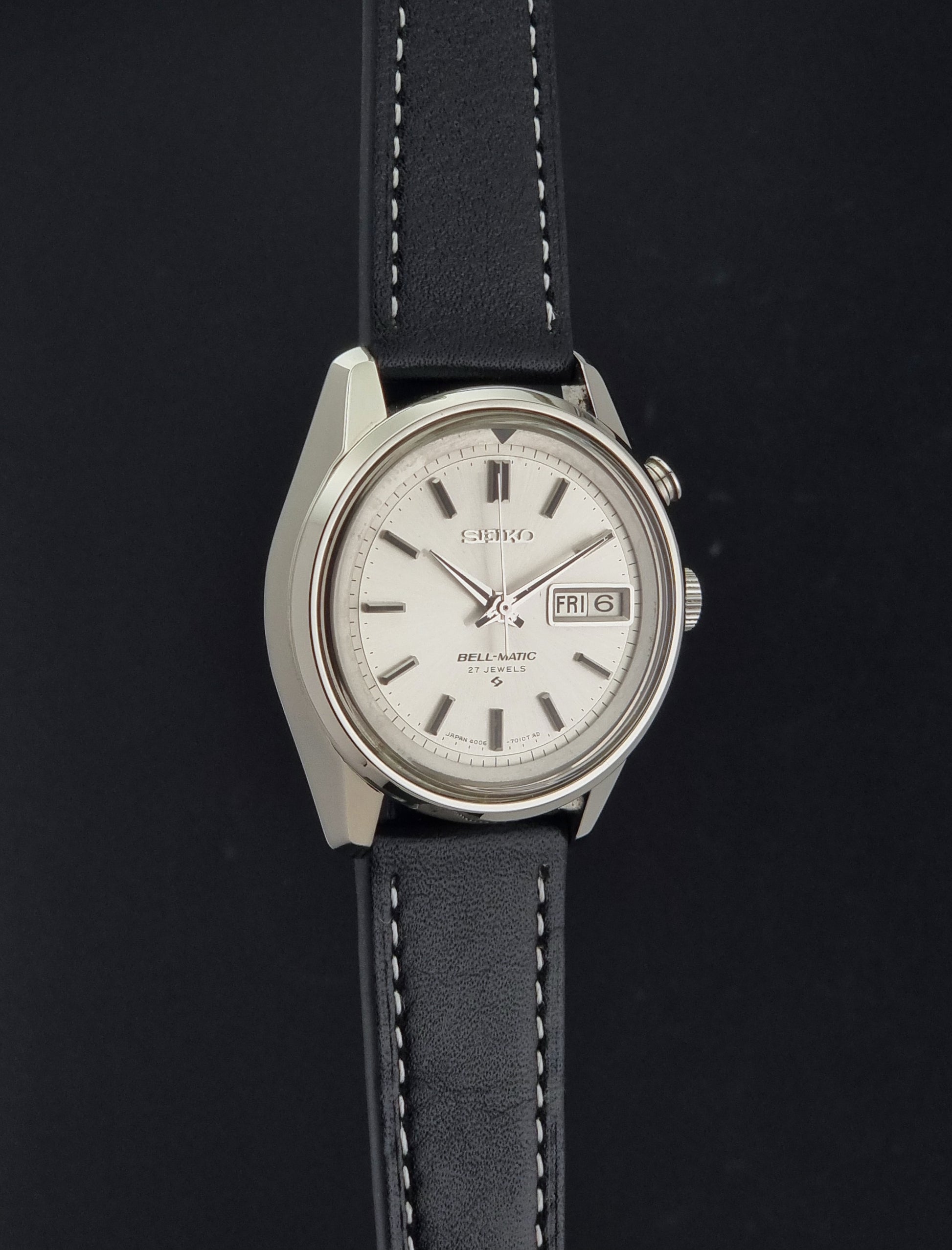 A Vintage Seiko  Bell Matic Alarm 4006-7010 watch with a silver-toned metal case, a white dial with a date window, and a black leather strap with white stitching, shown against a dark background