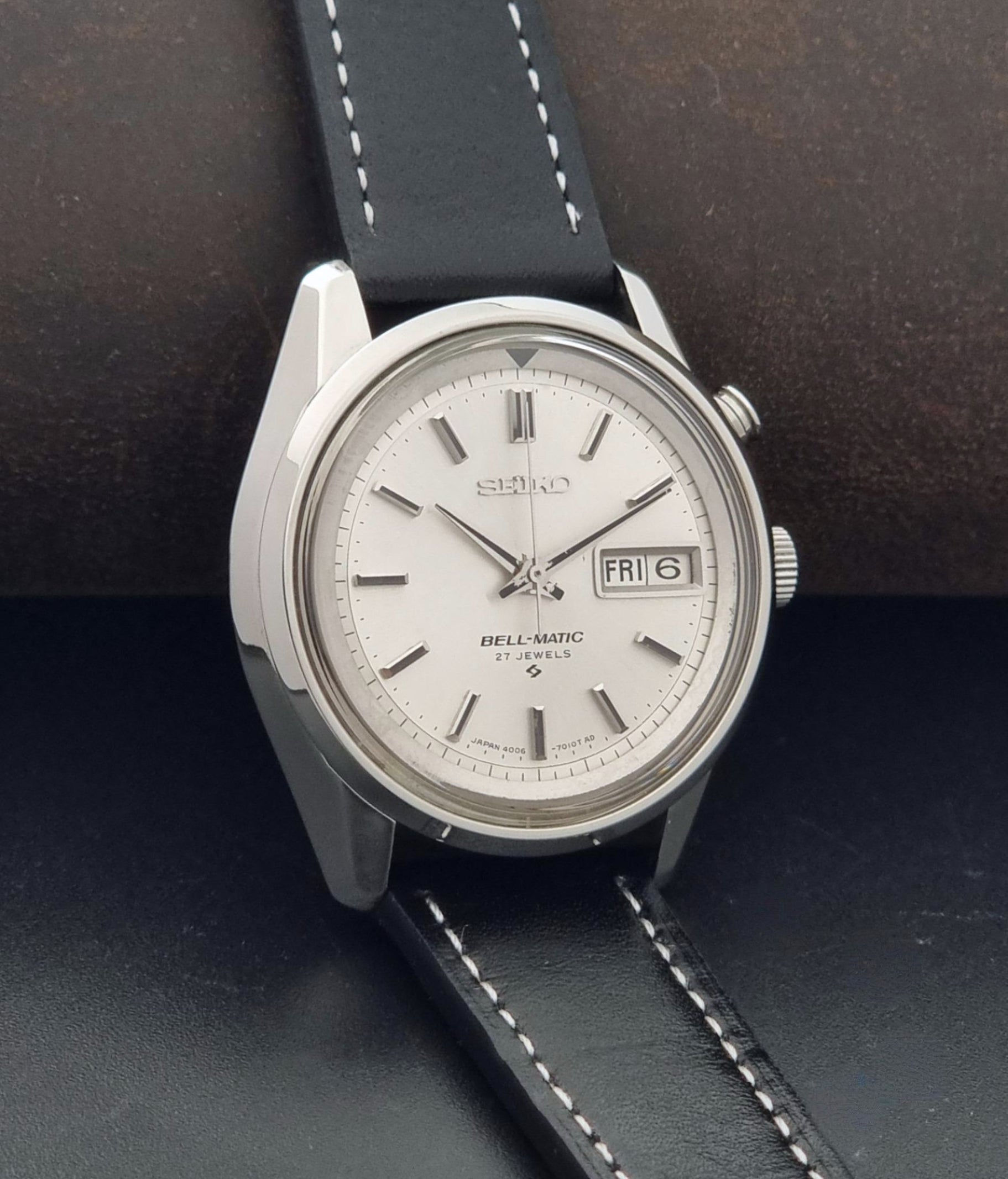A Vintage Seiko Wristwatch Bell Matic Alarm 4006-7010, white dial, and black leather strap with white stitching, resting on a dark textured surface.