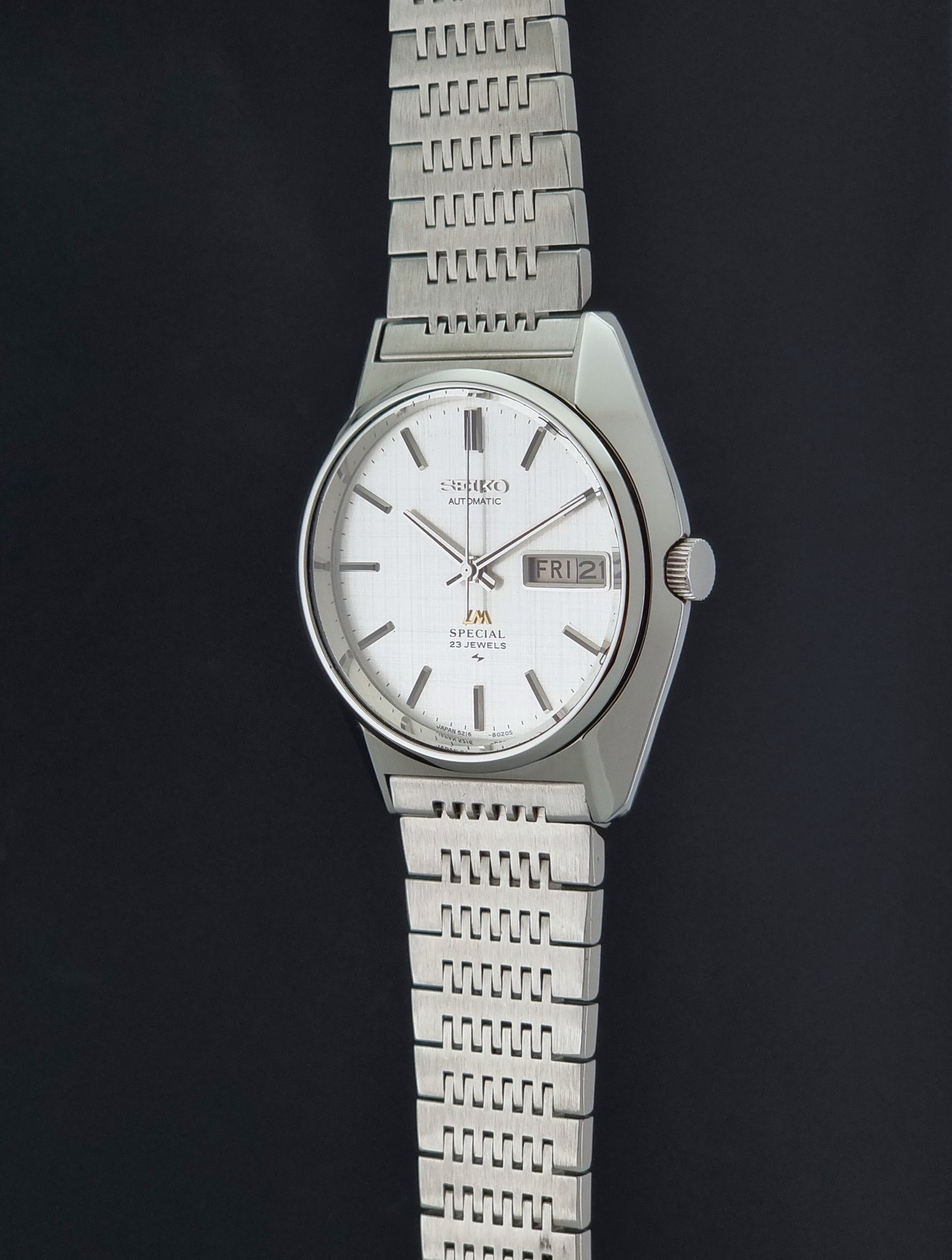 A Vintage Seiko LordMatic Special 5216-8020 Wristwatch with a silver-toned metal link bracelet and a white dial, shown against a dark background.