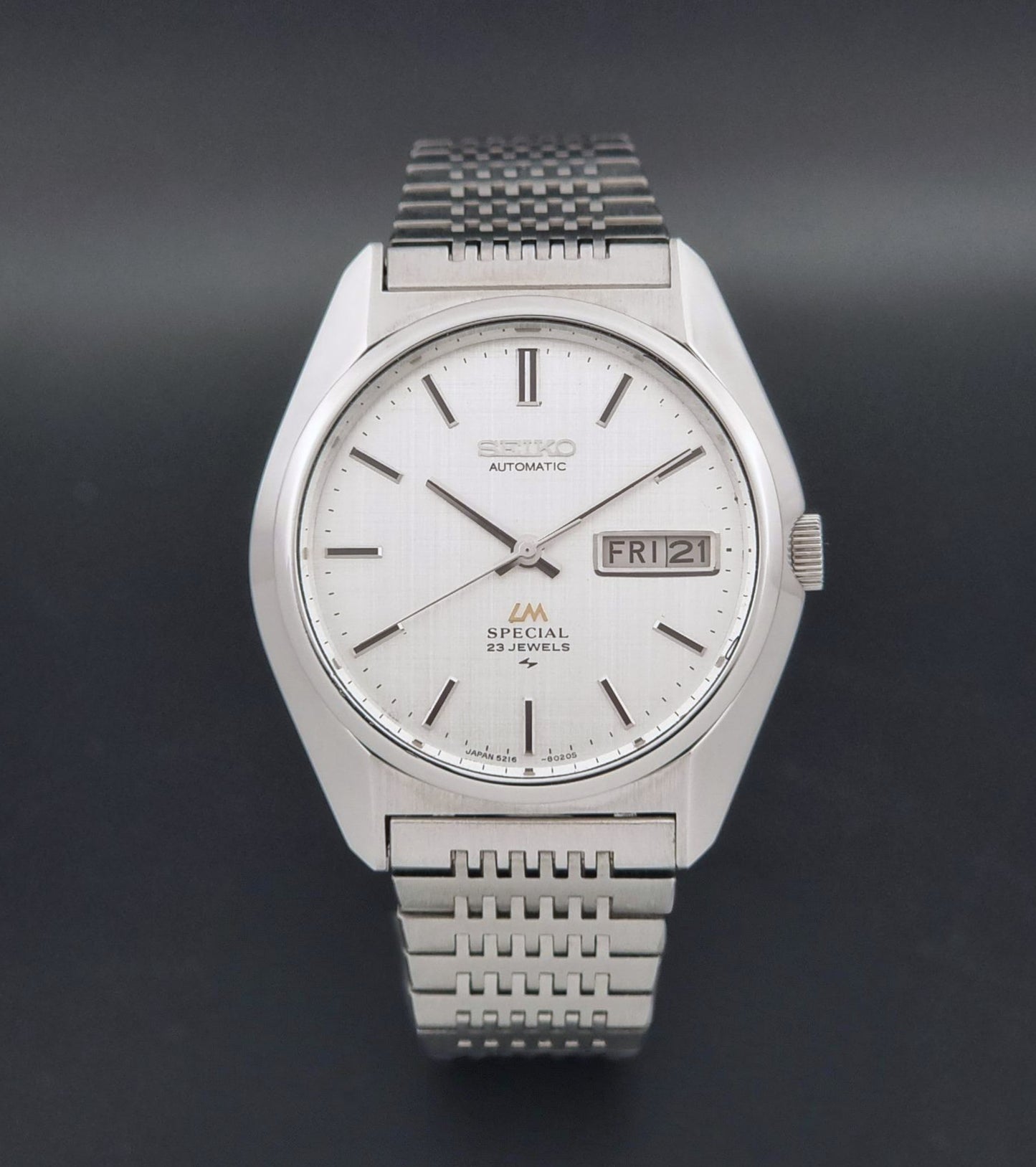 A Vintage Seiko LordMatic Special 5216-8020 Wristwatch featuring a white dial with black indices and a date window at 3 o'clock, set against a dark gray background.