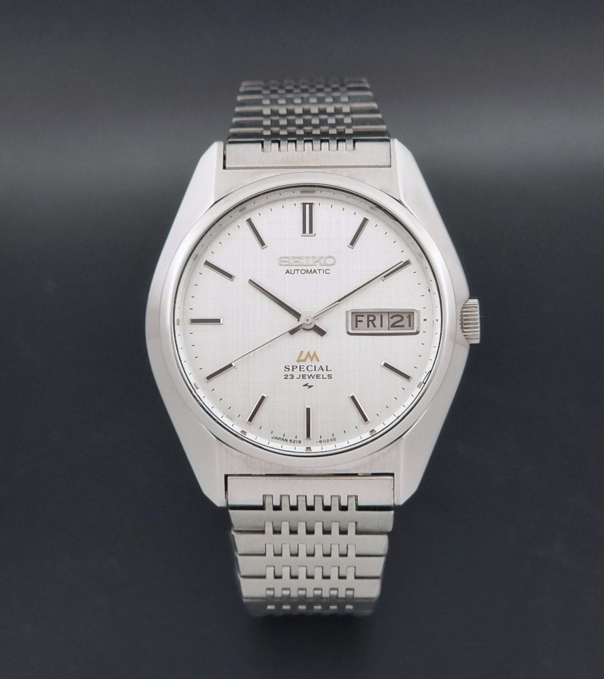 A Vintage Seiko LordMatic Special 5216-8020 Wristwatch featuring a white dial with black indices and a date window at 3 o'clock, set against a dark gray background.