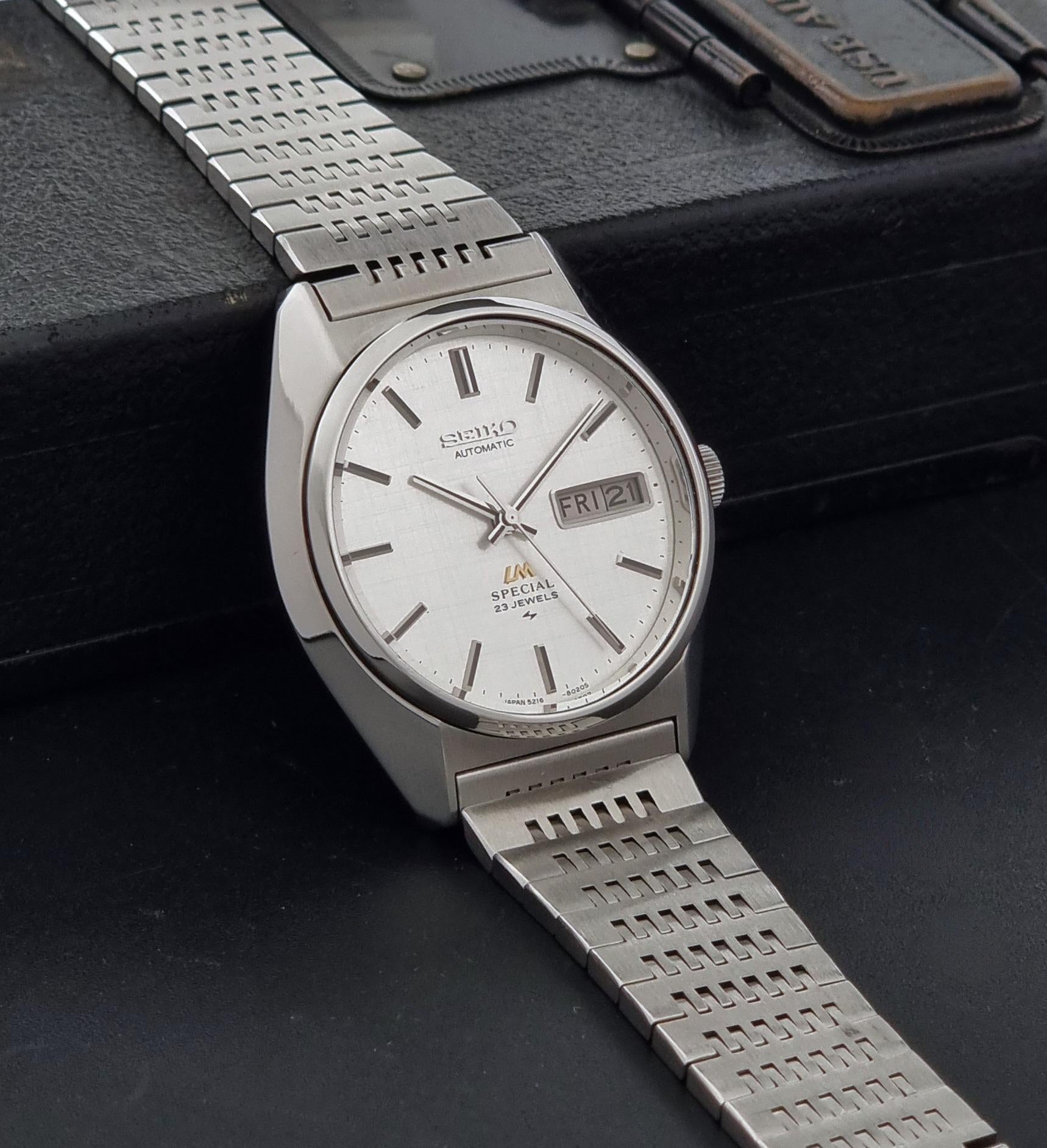 A Vintage Seiko LordMatic Special 5216-8020 Wristwatch with a silver-toned metal bracelet and a white dial, resting on a black textured surface with its original box partially visible in the background.