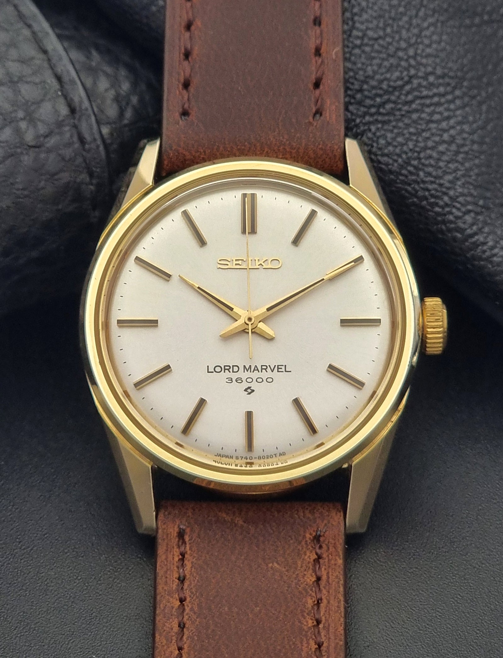 Vintage Seiko Lord Marvel 5740-8000 gold watch with a brown leather strap on a dark gray background close up dial