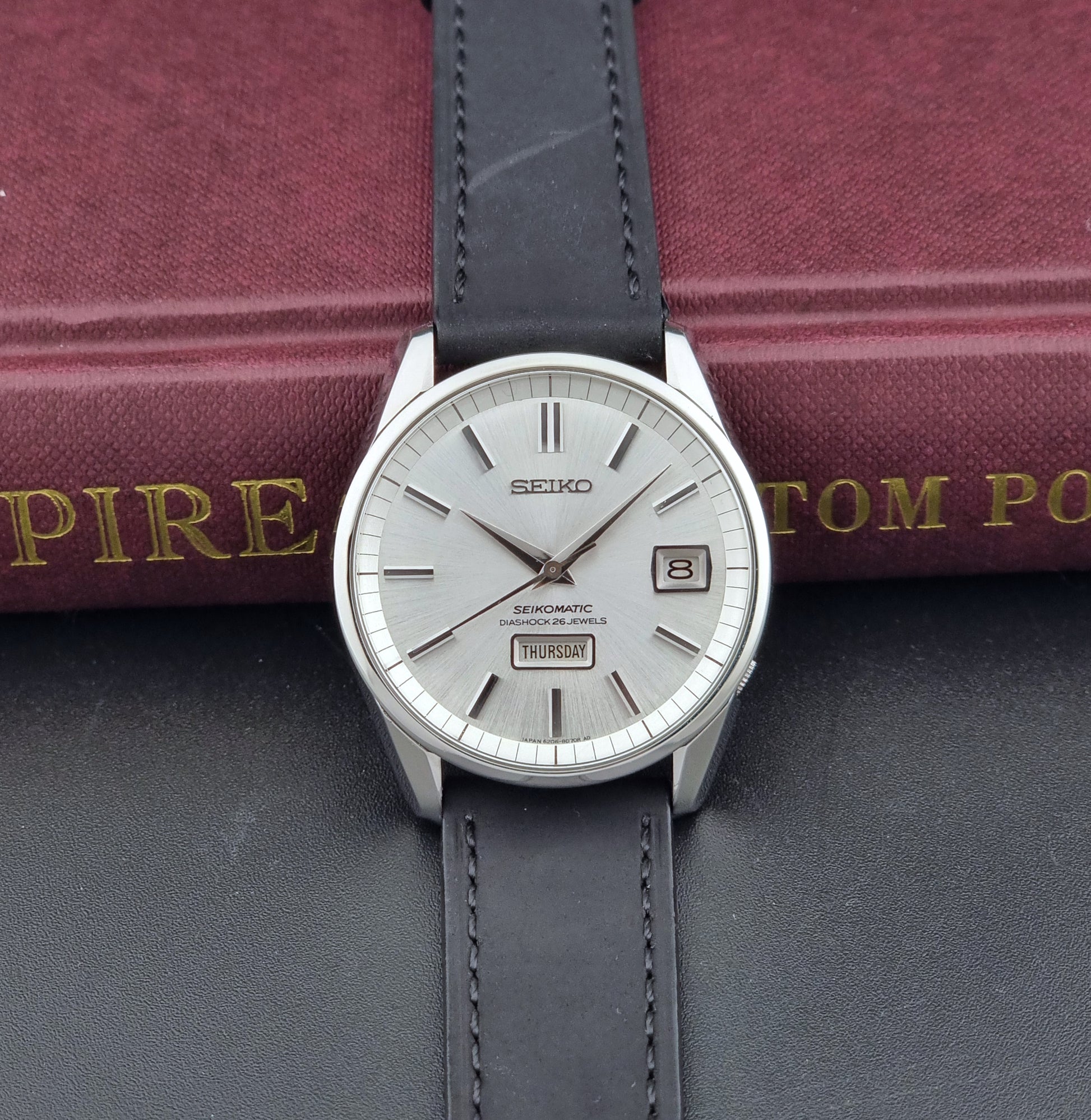 Seiko Seikomatic 6206-8040 Wristwatch with a silver dial and black leather strap on a dark surface with a book in the background.