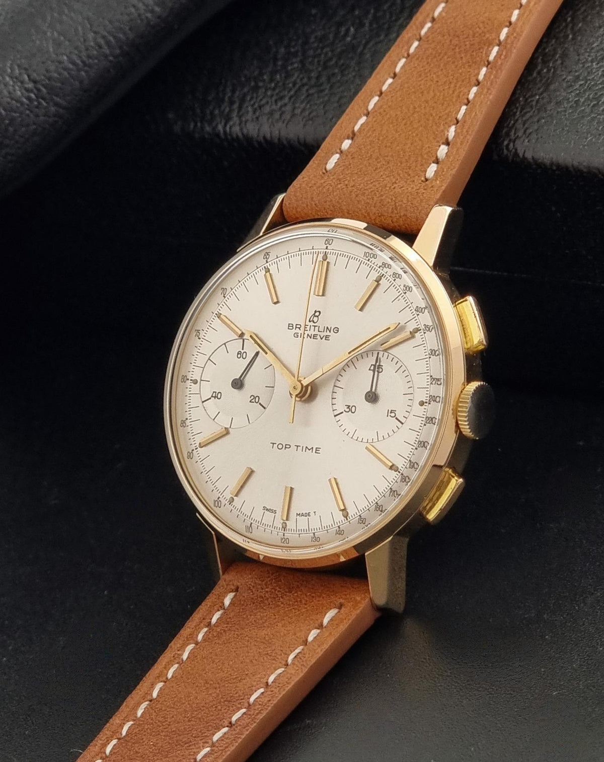 A vintage Breitling watch with a gold-tone case, white dial, and brown leather strap, photographed on a dark textured background.