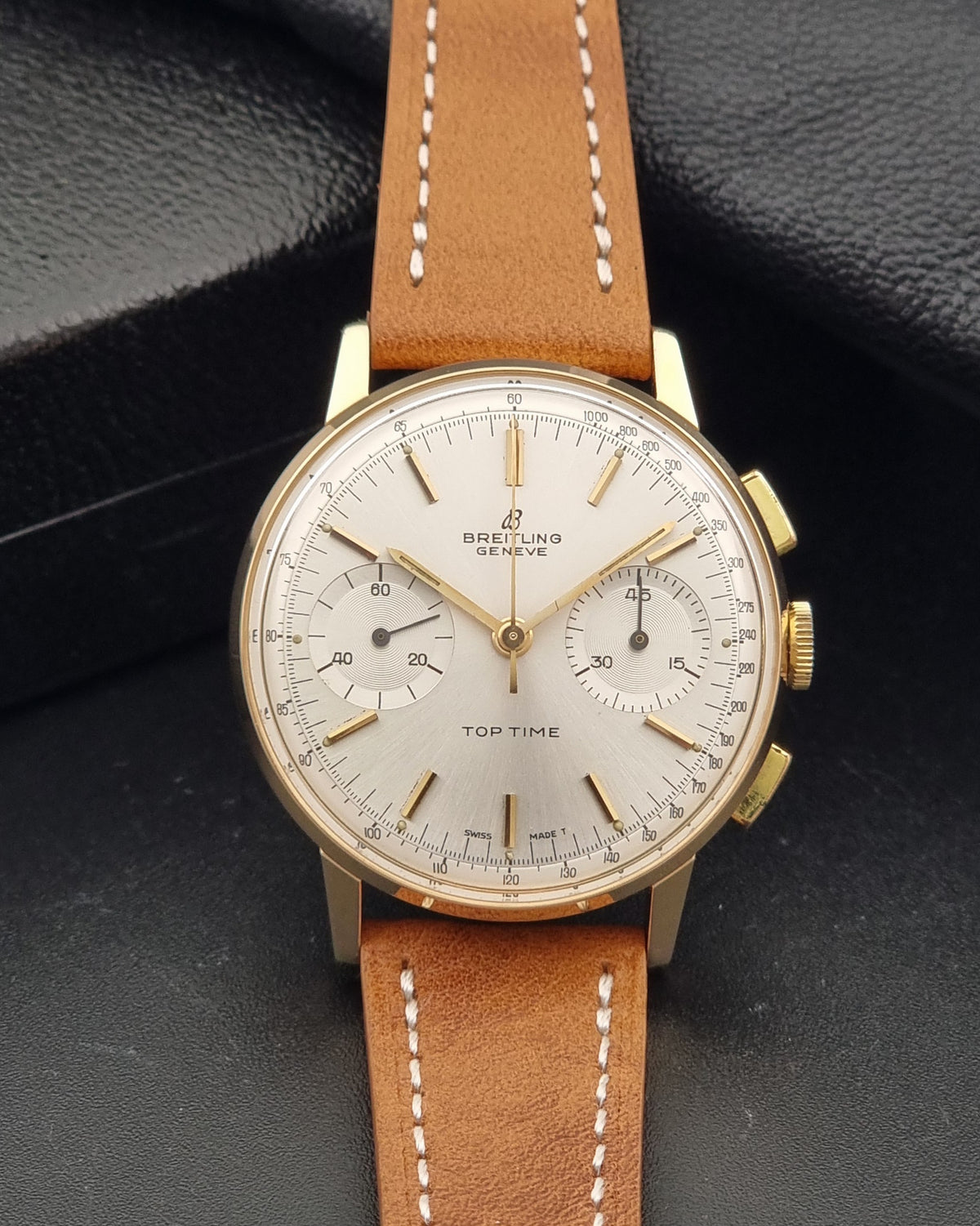 A Breitling Geneva Top Time watch with a gold-tone case, white dial, and a light brown leather strap with white stitching, displayed on a textured black background.