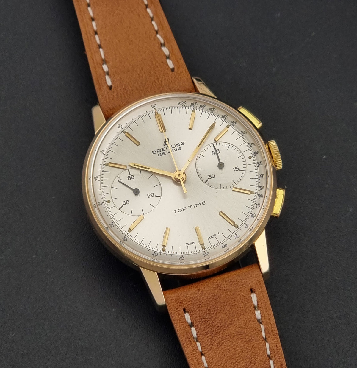 Vintage Breitling Top Time 2003 Chronograph watch with a gold-tone case, silver dial, and a brown leather strap with white stitching, photographed on a dark textured background.