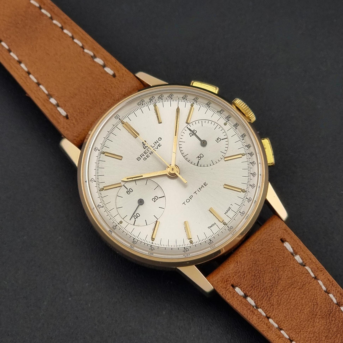 A vintage Breitling watch with a gold case, white dial, and brown leather strap, photographed on a dark textured background.