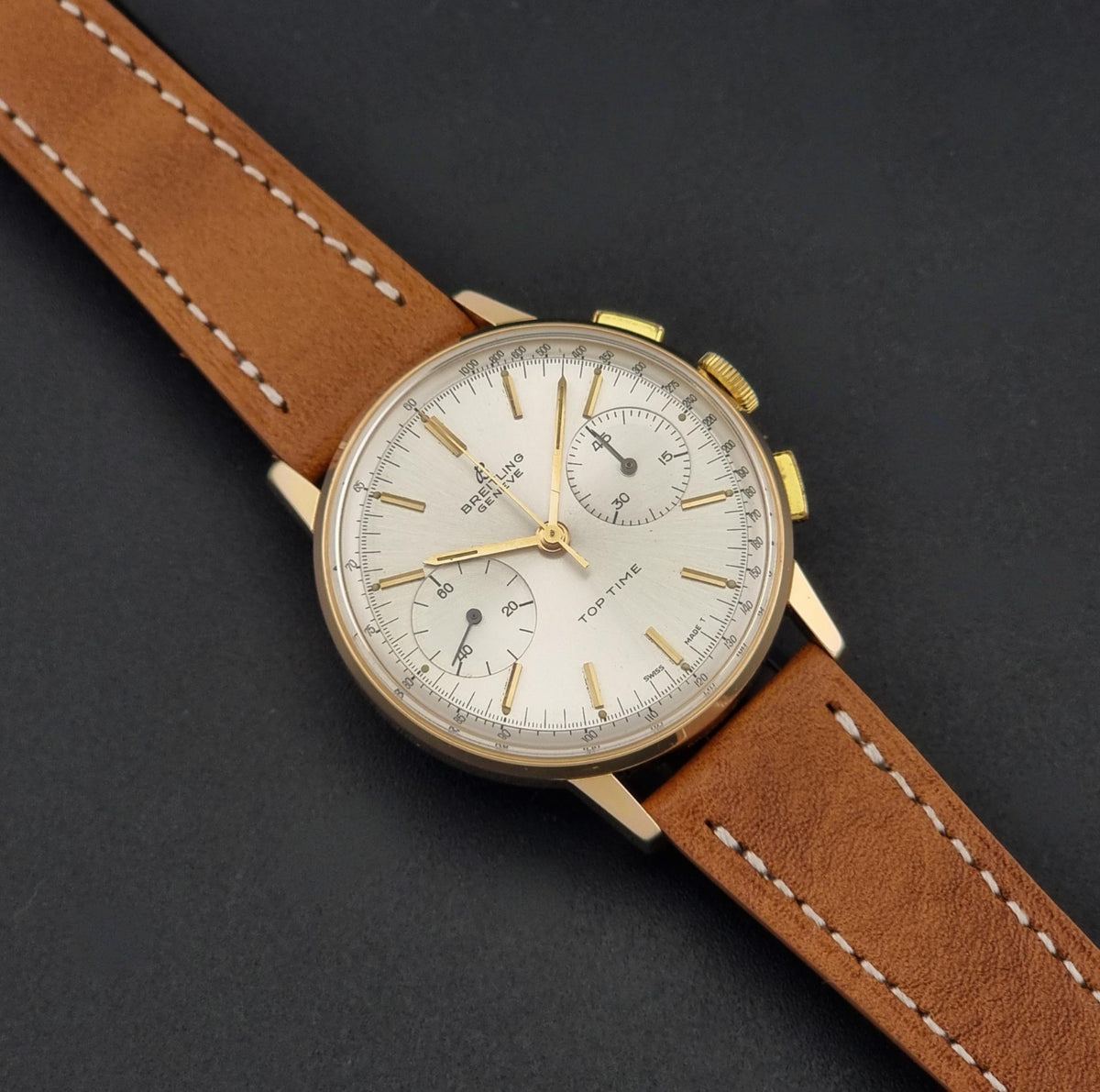 Vintage Breitling Top Time 2003 Chronograph watch with a round gold-tone case, white dial with three subdials, and a brown leather strap with white stitching, photographed on a dark textured background.