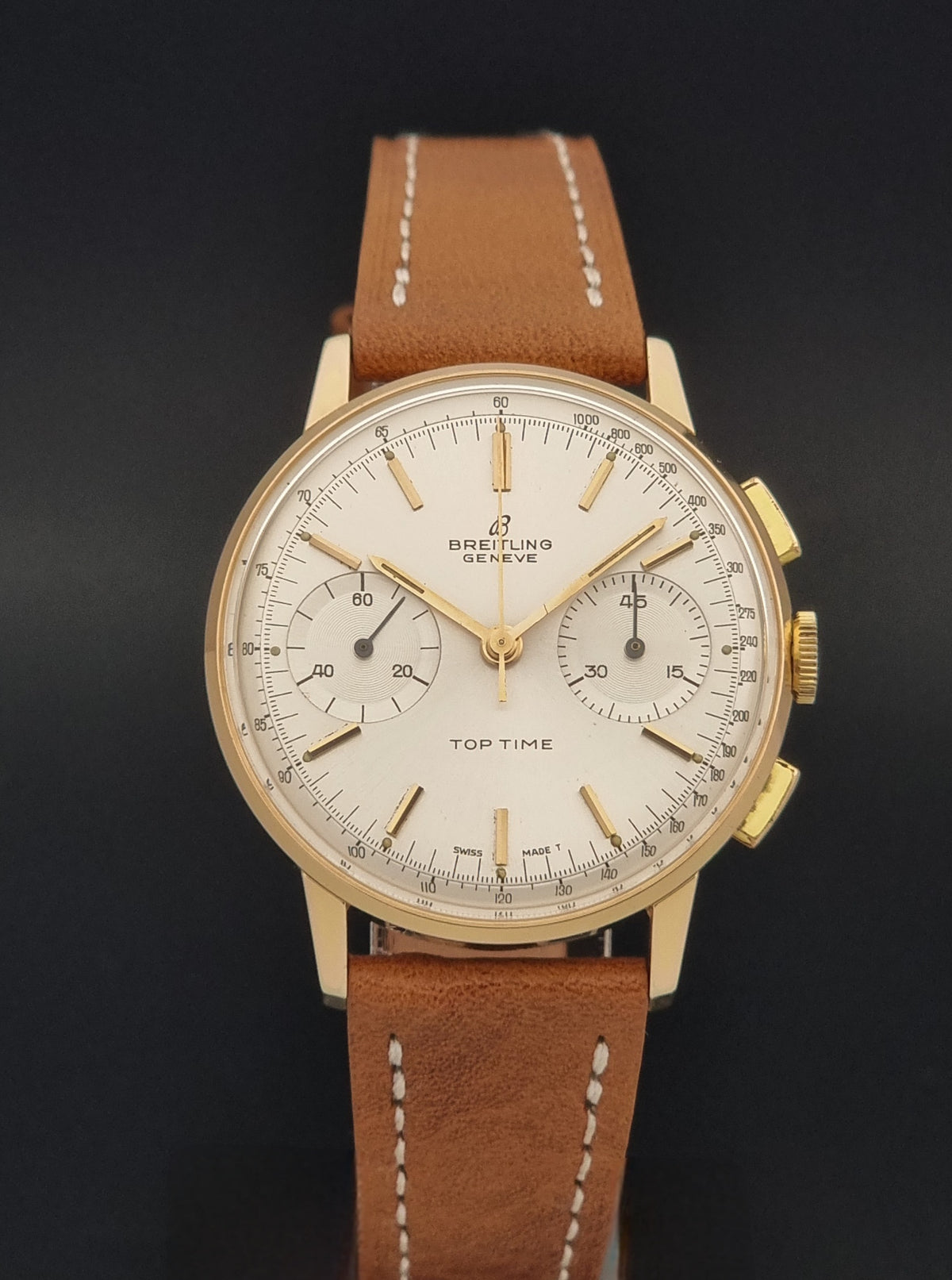 A Breitling Top Time wristwatch with a gold-tone case, white dial, and brown leather strap, shown against a dark background.