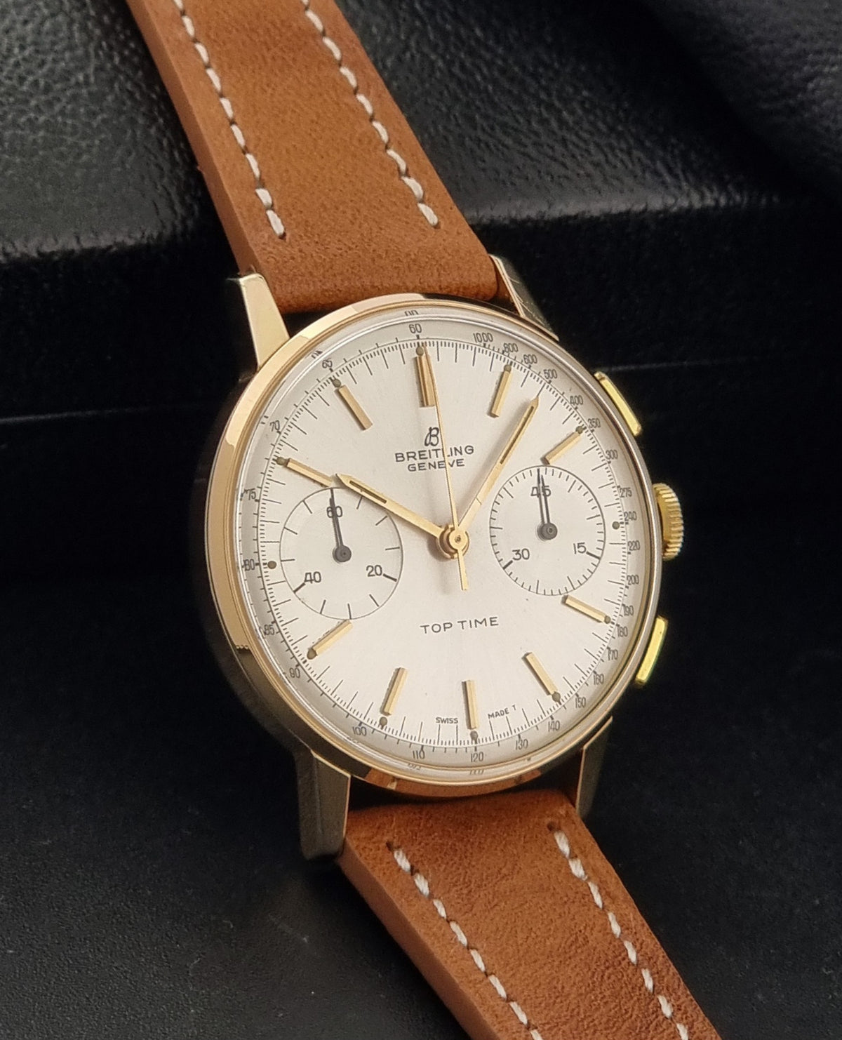 A vintage Breitling Top Time watch with a gold case, white dial, and a brown leather strap, displayed on a black textured background.