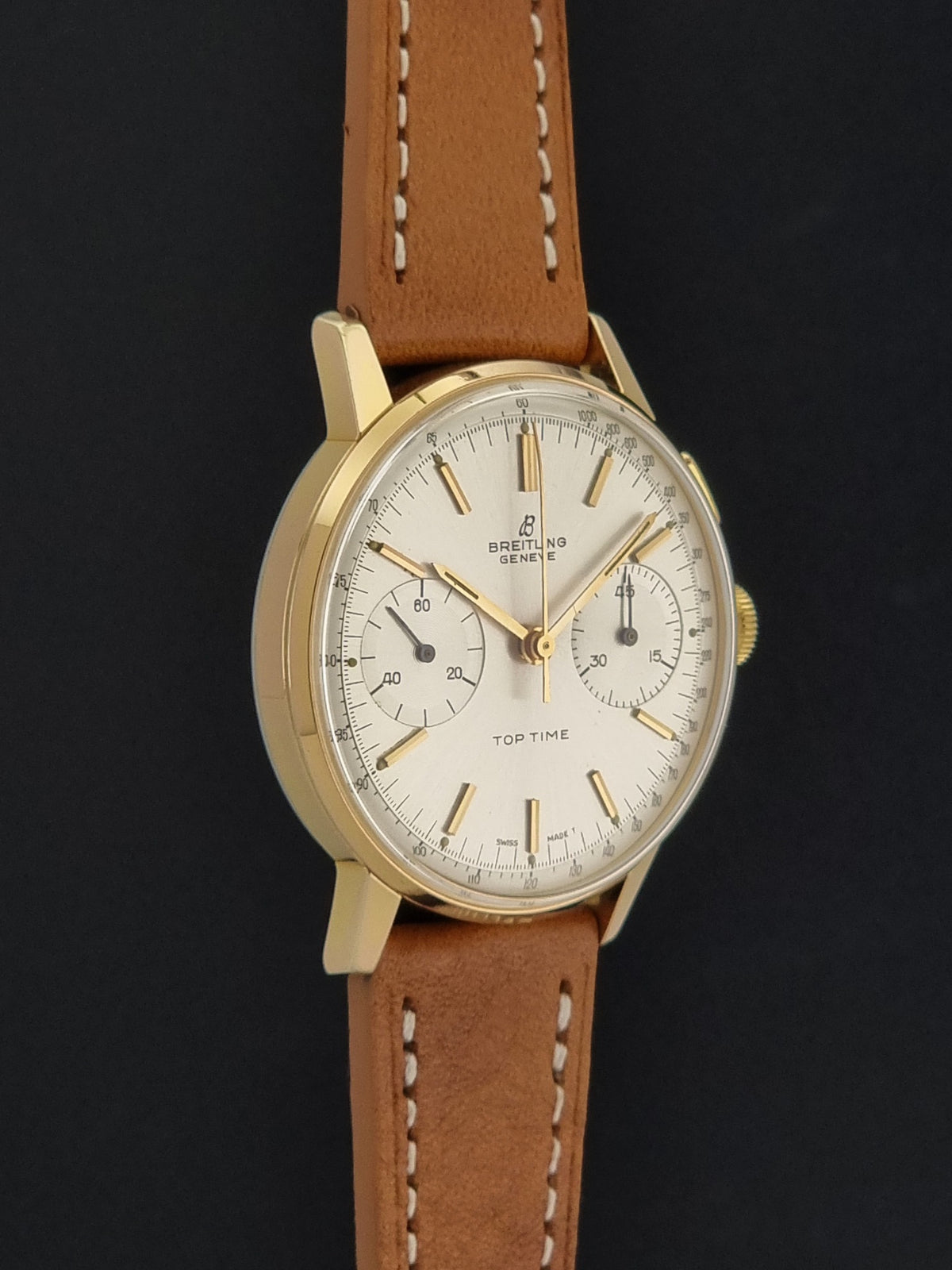 A vintage Breitling wristwatch with a gold-tone case, cream-colored dial, and a brown leather strap with white stitching, set against a dark background.