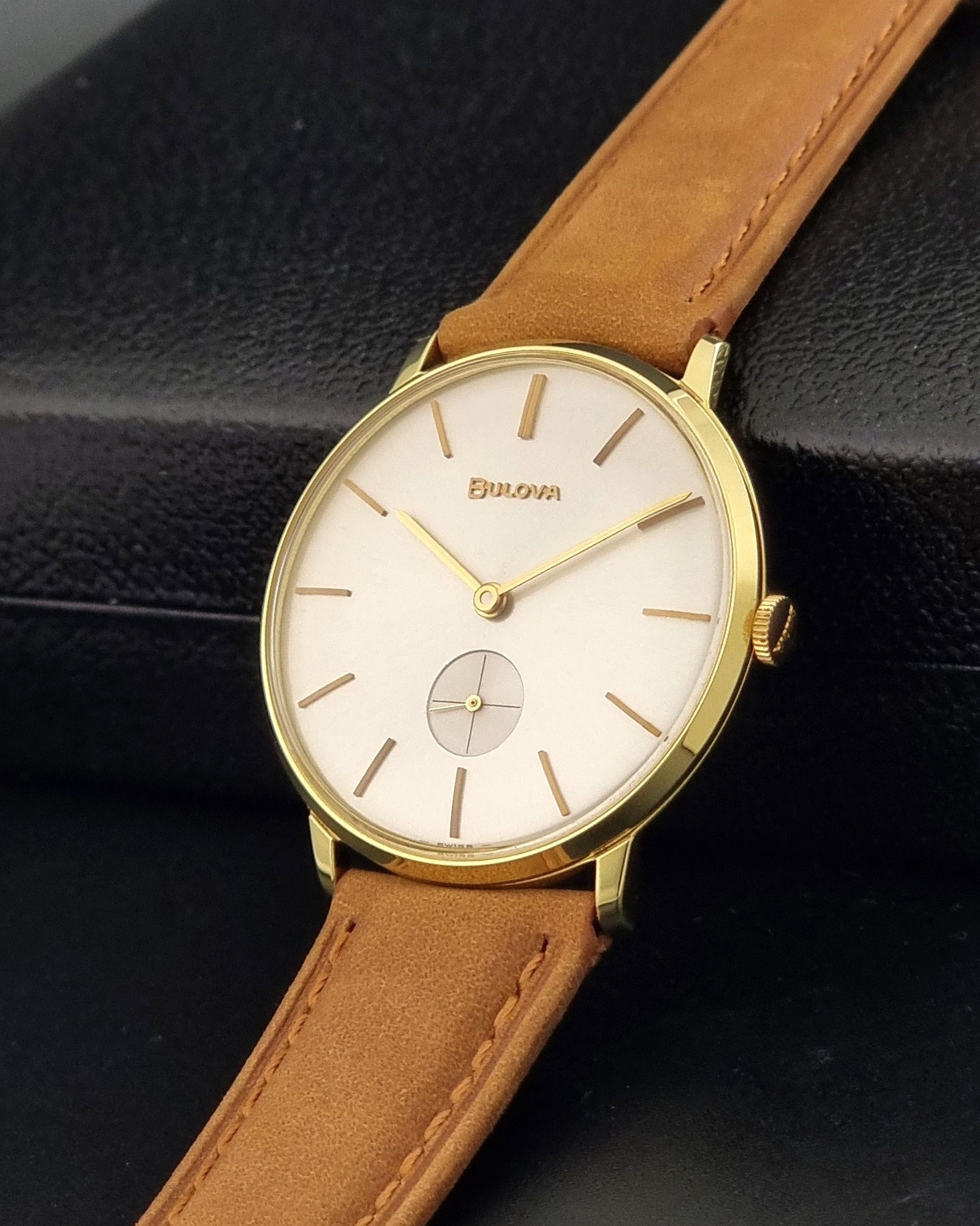 Vintage Bulova wristwatch 531  watch with a gold case, white dial, and a tan leather strap on a dark background crown view