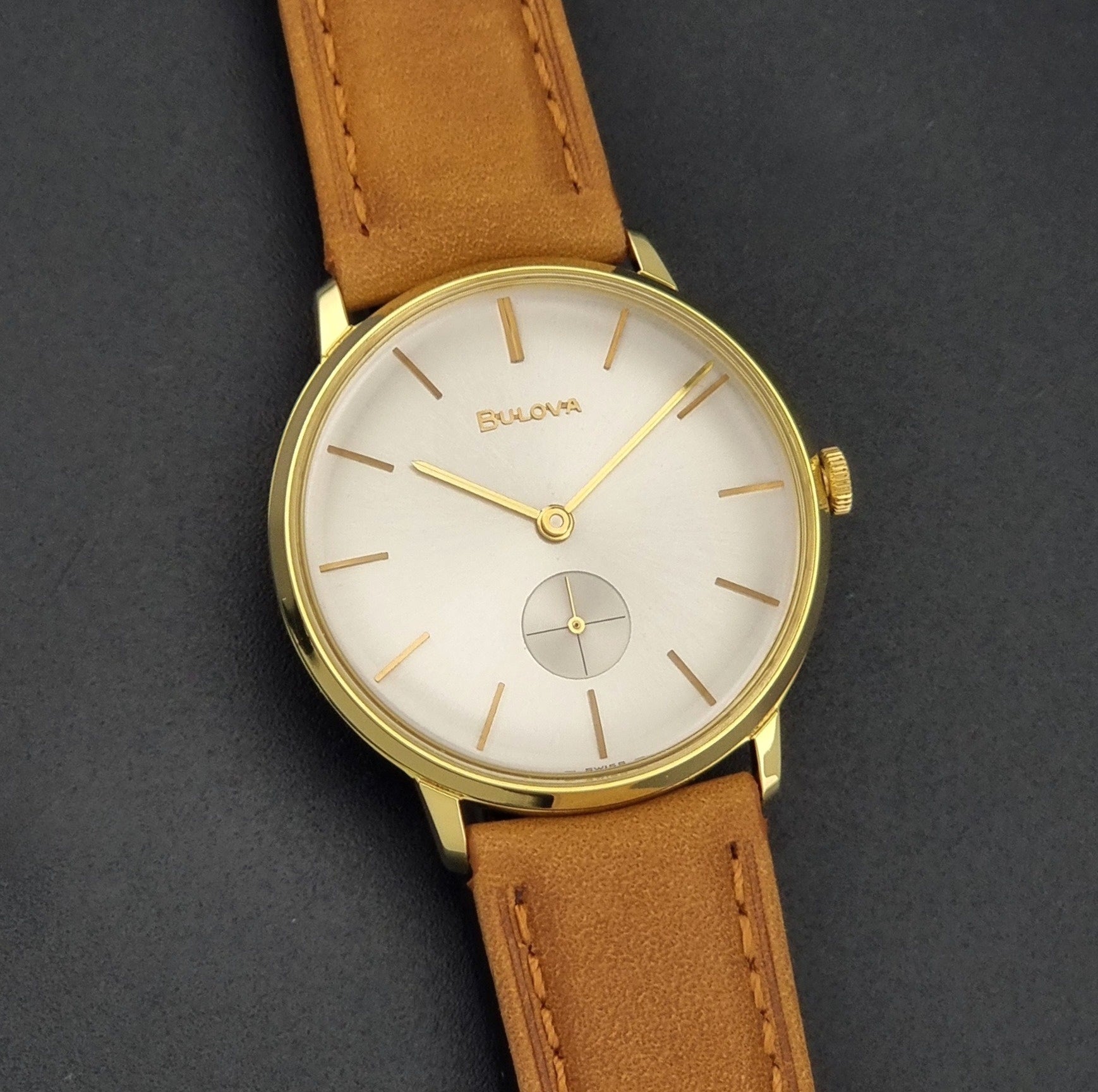 Vintage Bulova wristwatch 531  watch with a gold case, white dial, and a tan leather strap on a dark background front view
