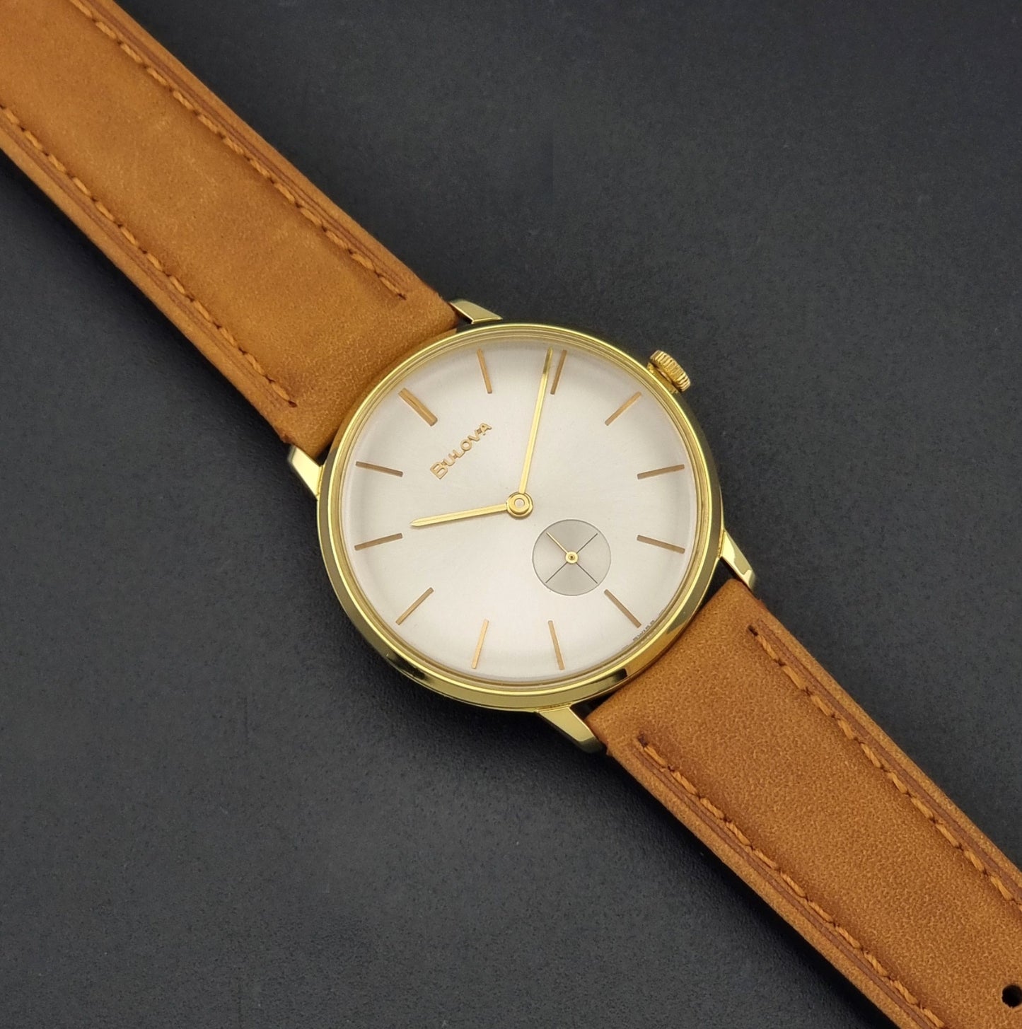 Vintage Bulova wristwatch 531  watch with a gold case, white dial, and a tan leather strap on a dark background dial