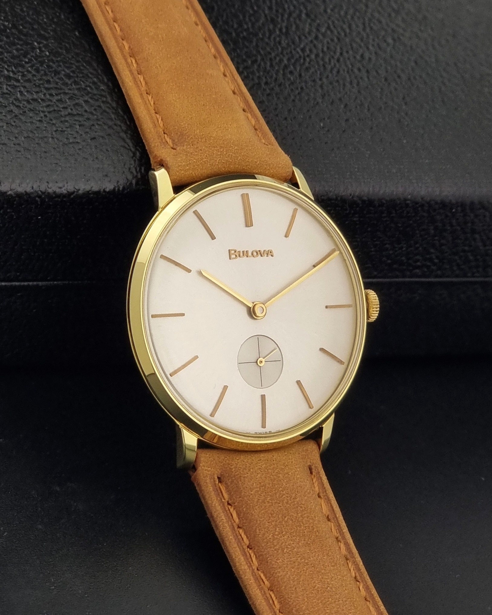 Vintage Bulova wristwatch 531  watch with a gold case, white dial, and a tan leather strap on a dark background side view