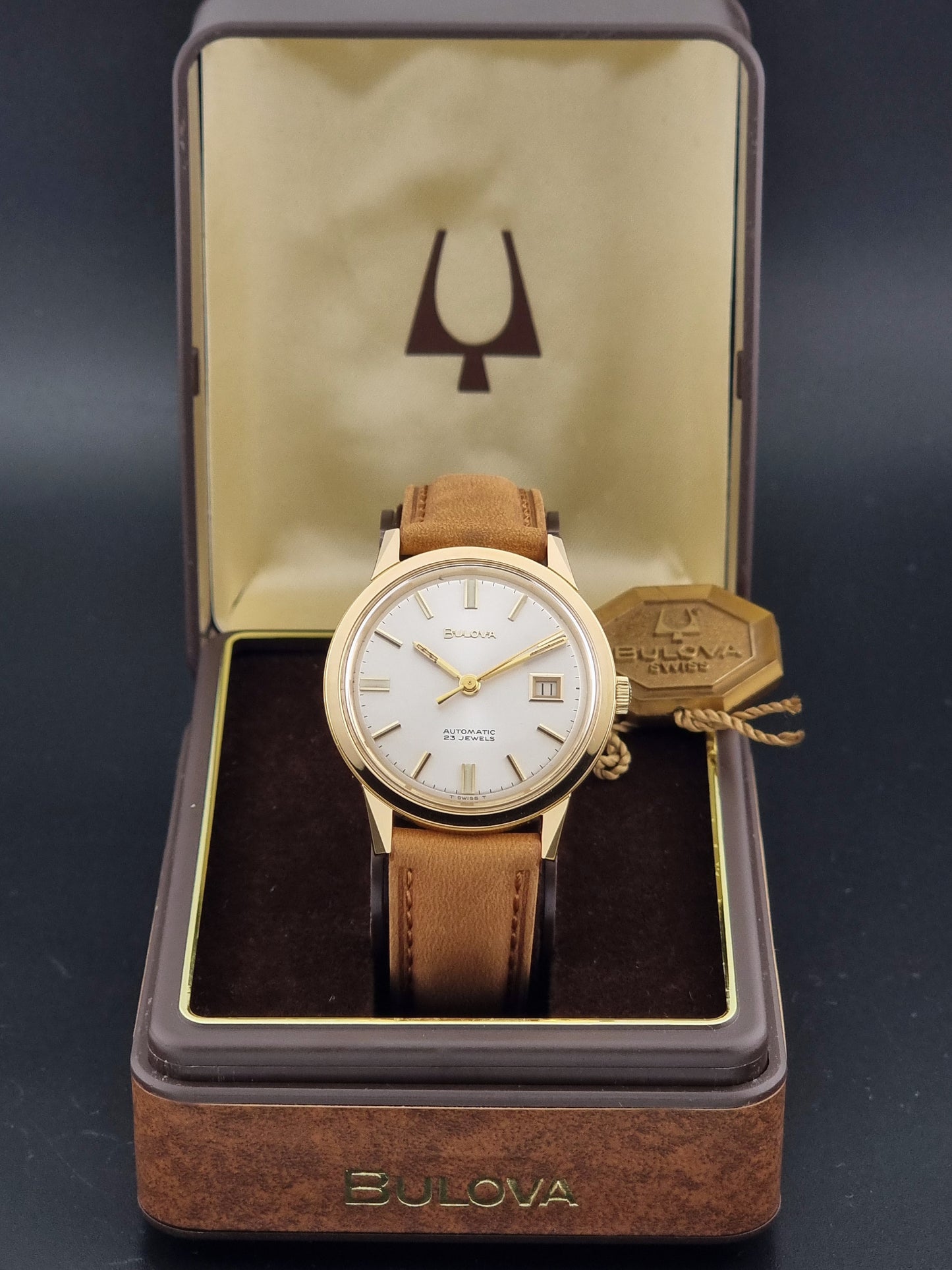 Vintage Bulova wristwatch 576-1  watch with a gold case, white dial, and a tan leather strap on a dark background box view