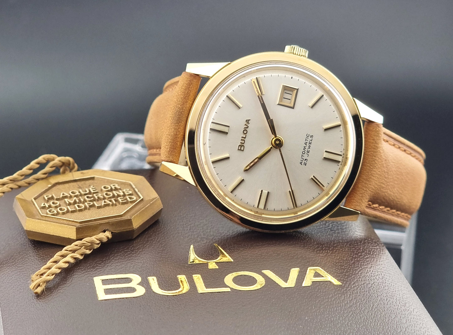 Vintage Bulova wristwatch 576-1  watch with a gold case, white dial, and a tan leather strap on a dark background dial view on box
