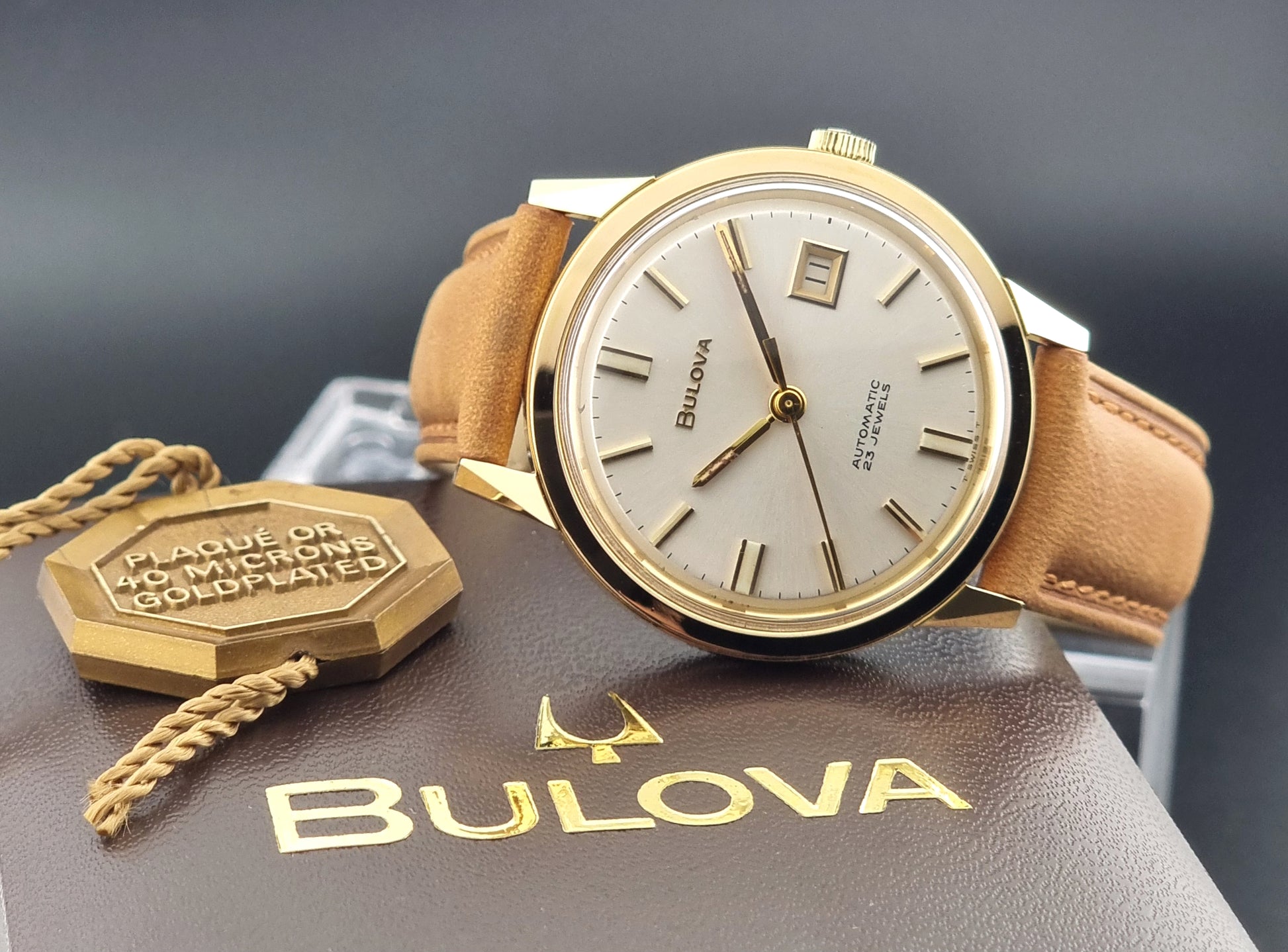 Vintage Bulova wristwatch 576-1  watch with a gold case, white dial, and a tan leather strap on a dark background dial view on box