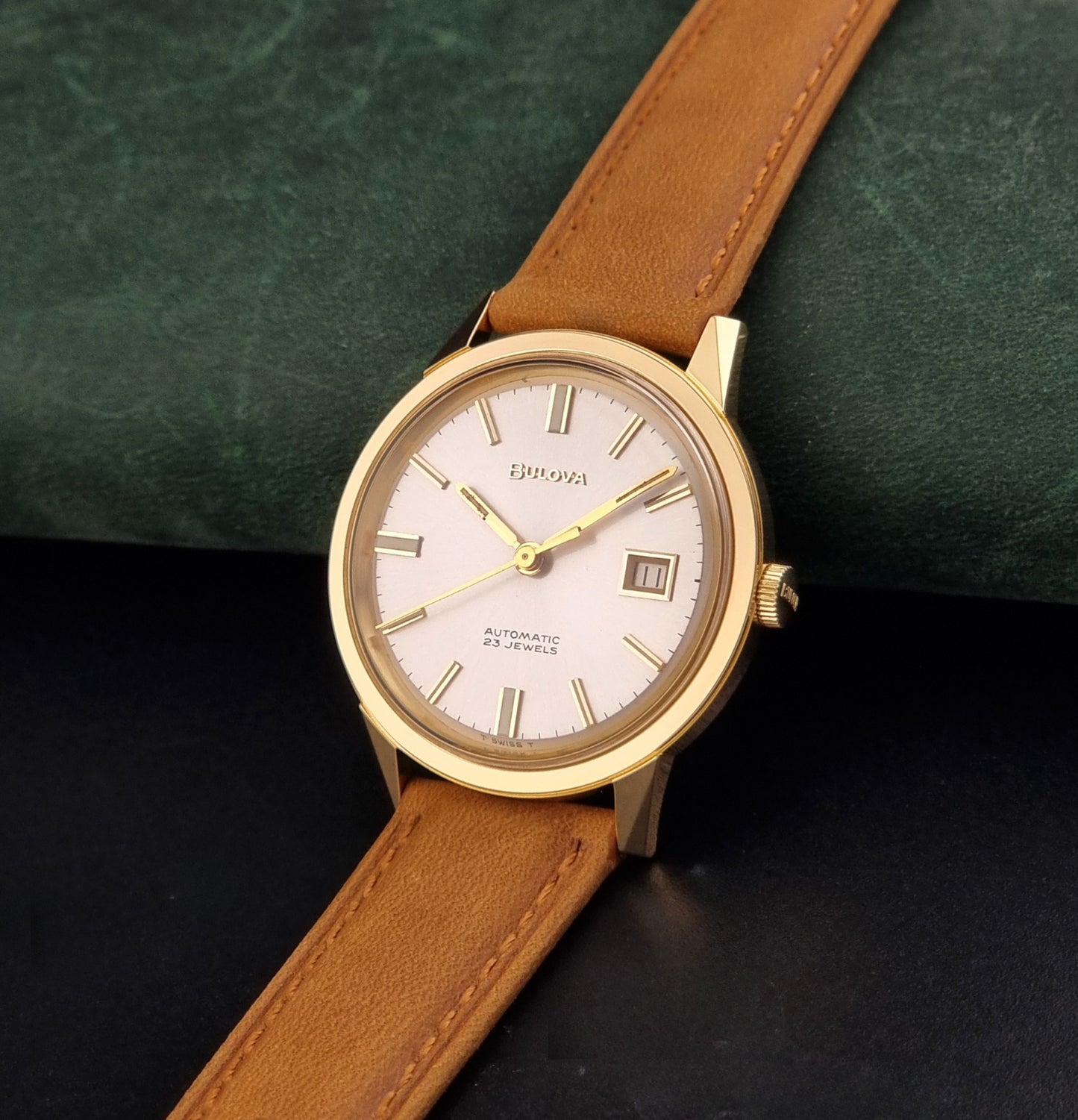 Vintage Bulova wristwatch 576-1  watch with a gold case, white dial, and a tan leather strap on a dark background crown view
