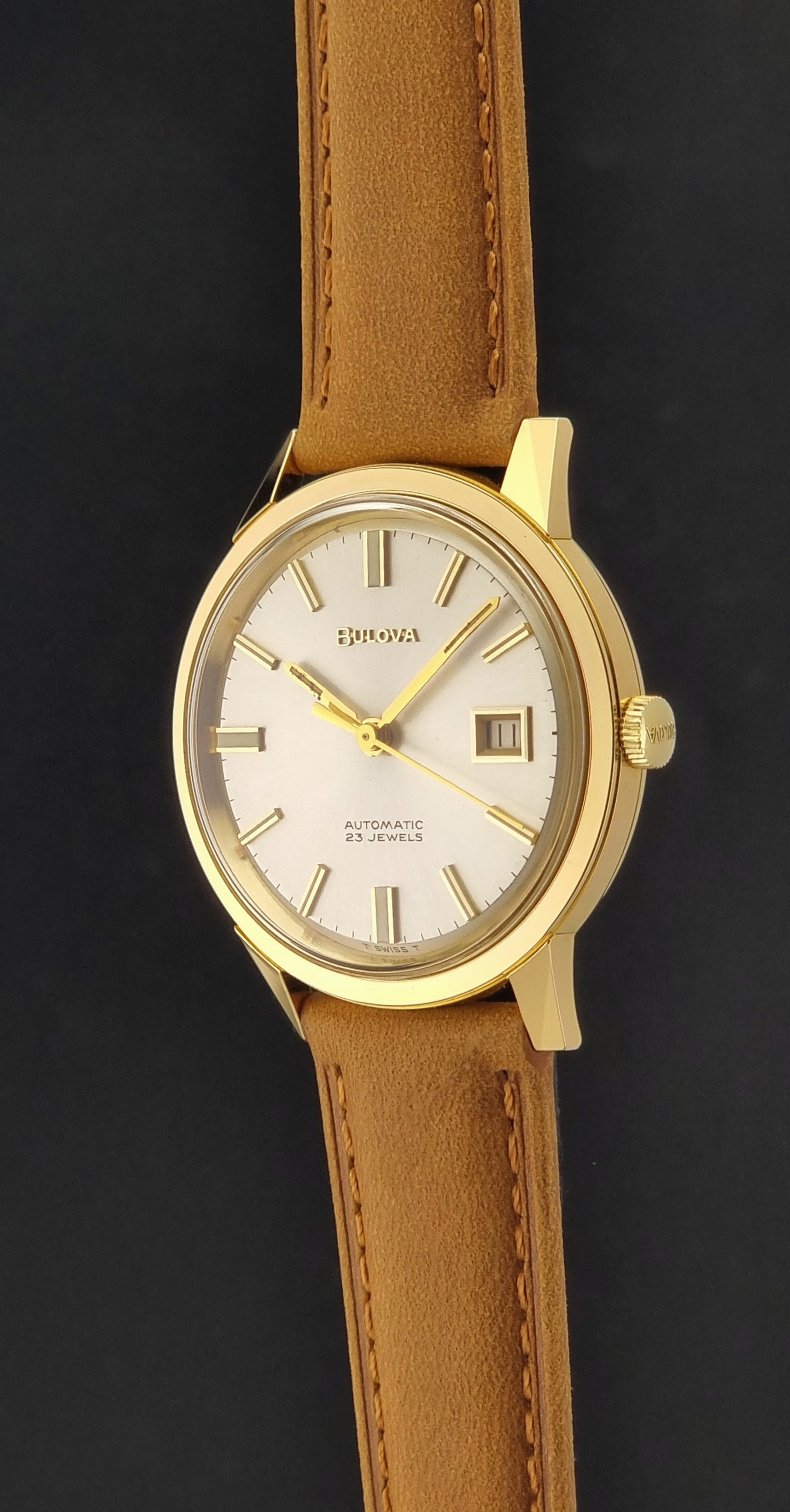 Vintage Bulova wristwatch 576-1  watch with a gold case, white dial, and a tan leather strap on a dark background crown profile view