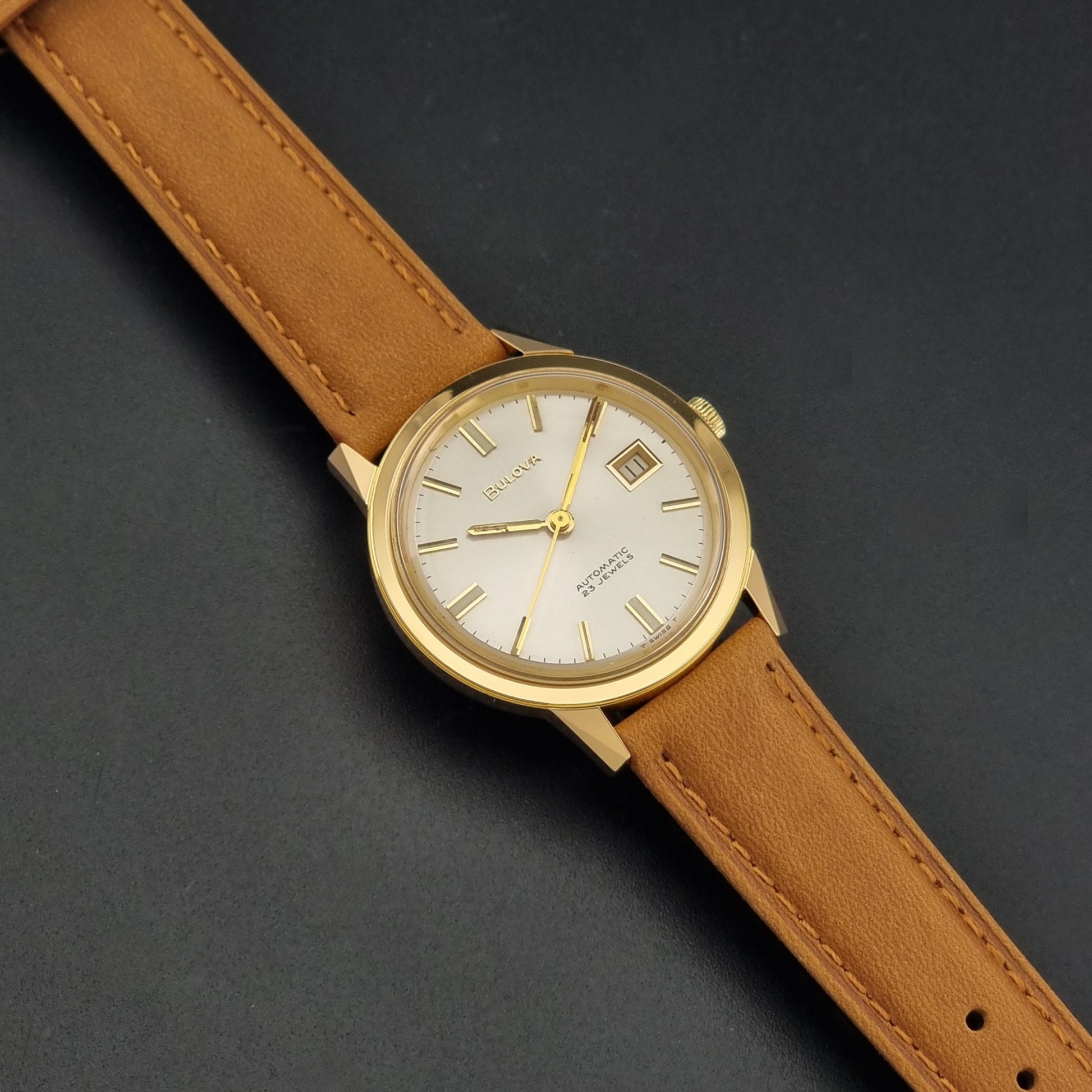 Vintage Bulova wristwatch 576-1  watch with a gold case, white dial, and a tan leather strap on a dark background dial