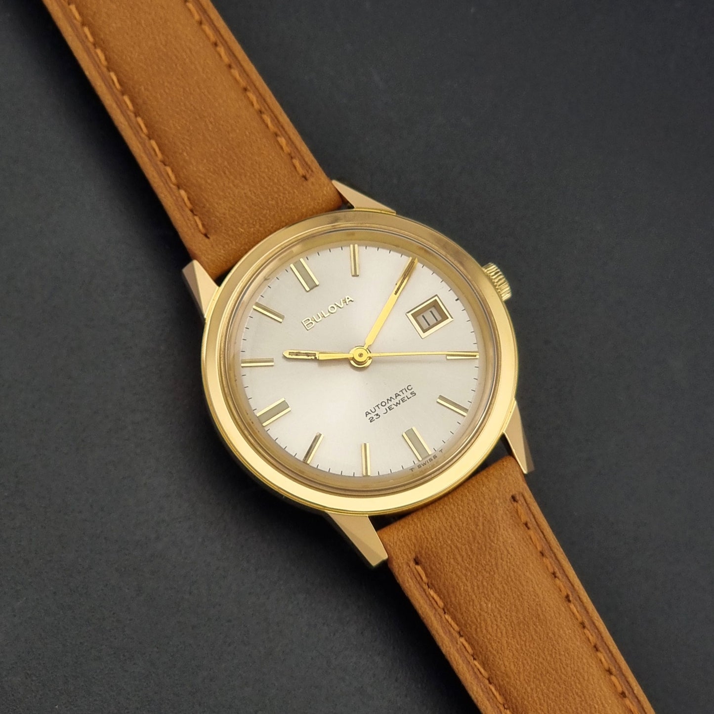 Vintage Bulova wristwatch 576-1  watch with a gold case, white dial, and a tan leather strap on a dark background front view