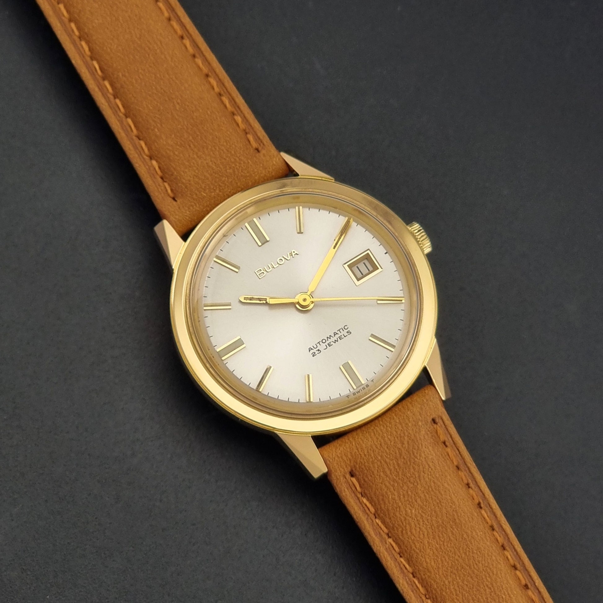 Vintage Bulova wristwatch 576-1  watch with a gold case, white dial, and a tan leather strap on a dark background front view