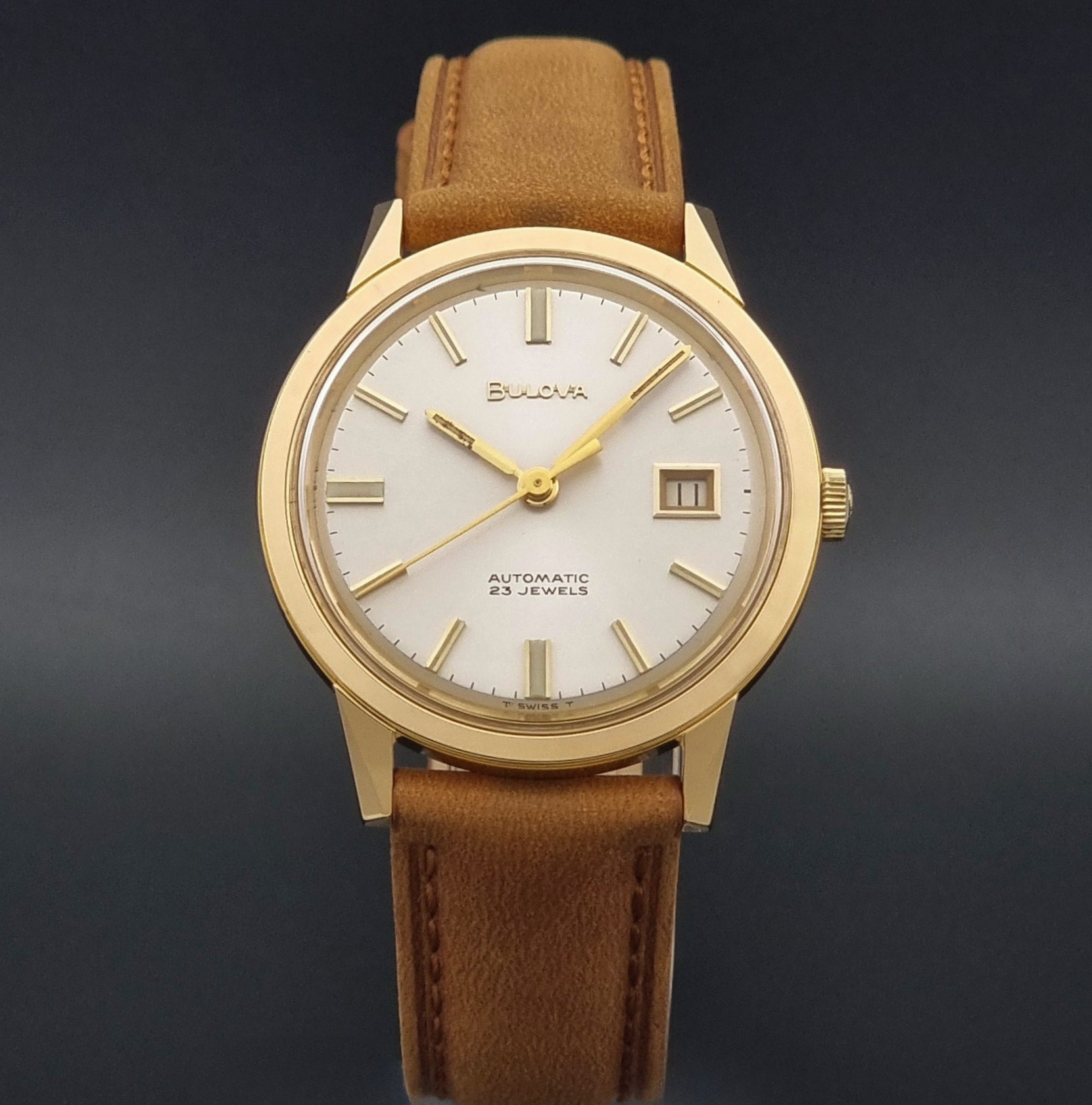 Vintage Bulova wristwatch 576-1  watch with a gold case, white dial, and a tan leather strap on a dark background close up dial view