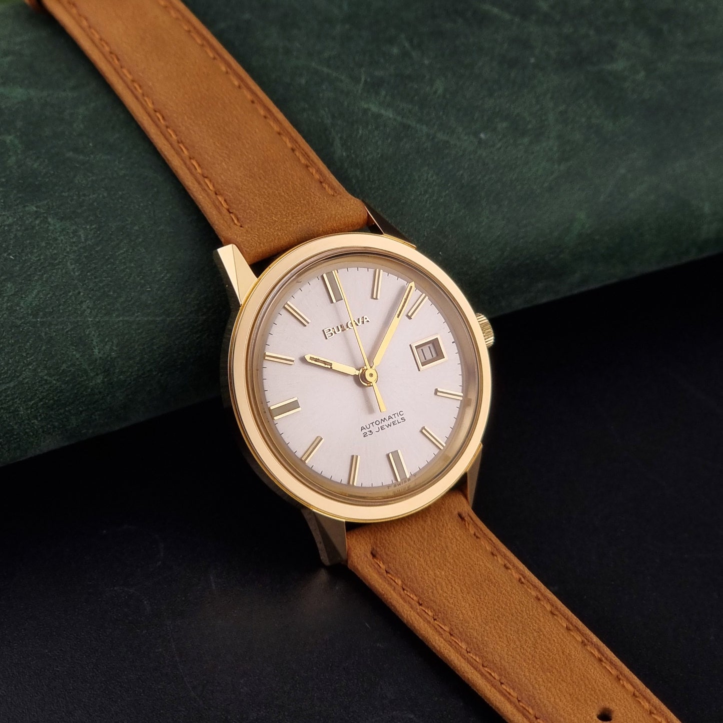 Vintage Bulova wristwatch 576-1  watch with a gold case, white dial, and a tan leather strap on a dark background side view