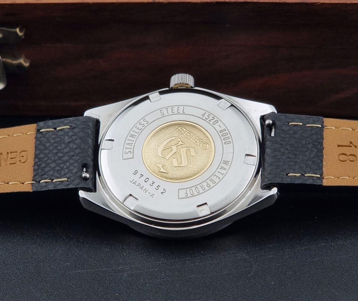 A vintage Grand Seiko watch 4520-8000 case back, with gold medallion, a stainless steel case and black leather strap on a dark background