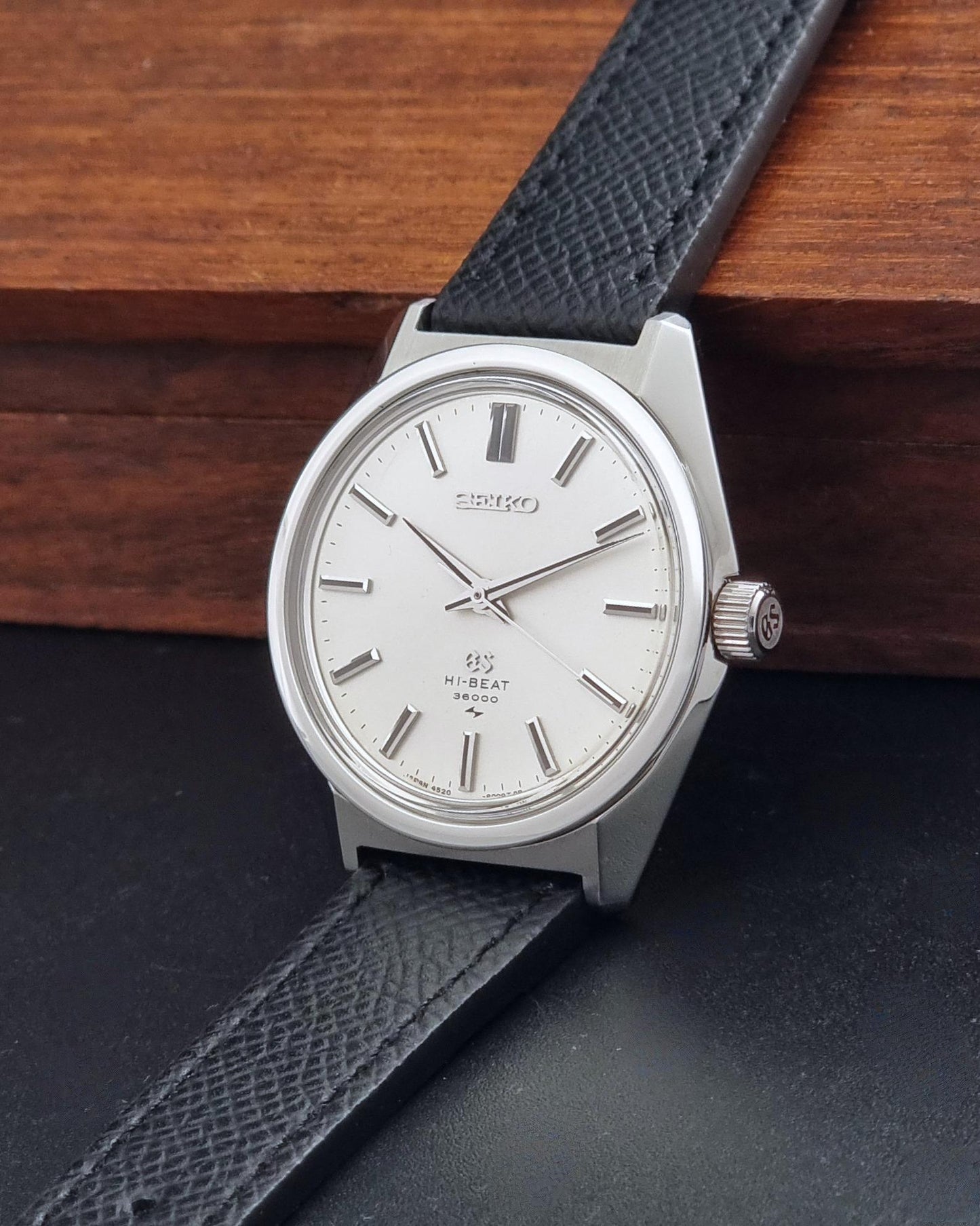 A vintage Grand Seiko watch 4520-8000 with a stainless steel case and black leather strap on a dark background looking at the crown resting on a wooden box