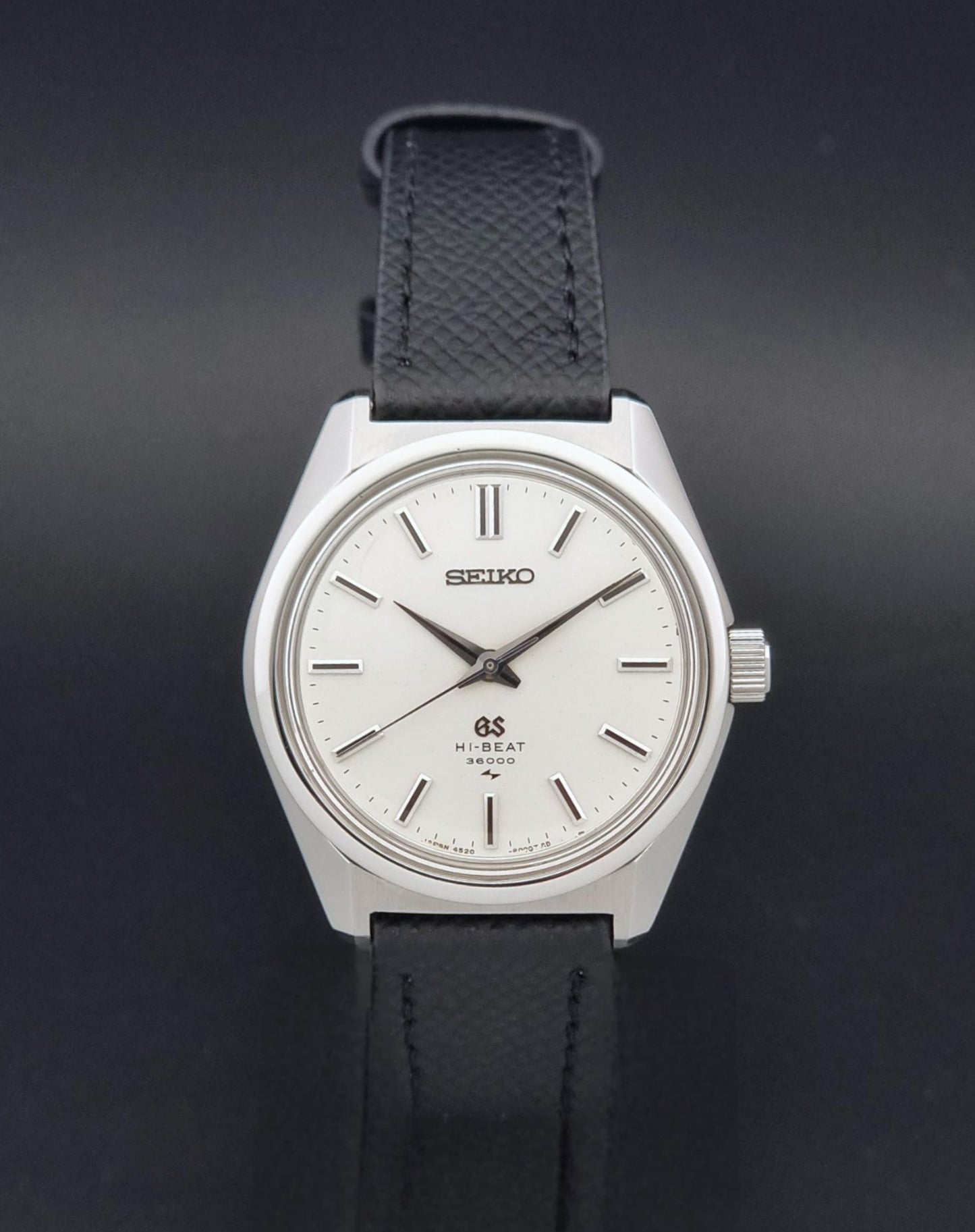 A vintage Grand Seiko watch 4520-8000 with a stainless steel case and black leather strap on a dark background viewing the sunburst dial