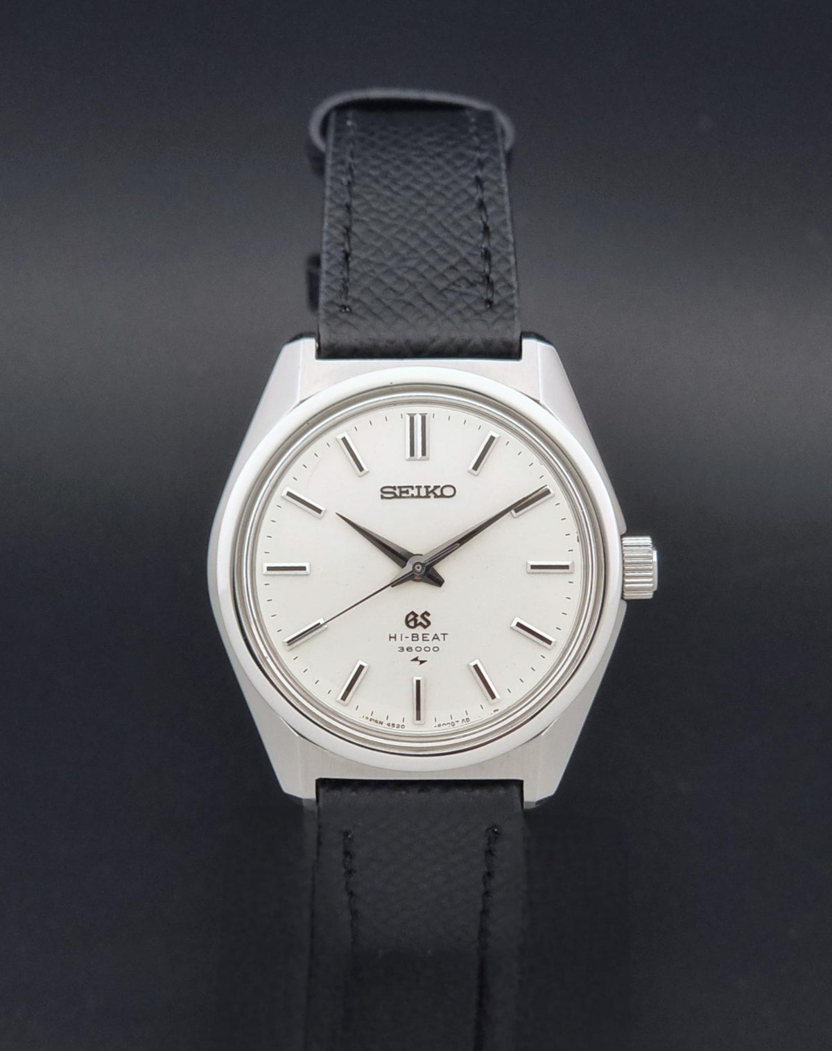 A vintage Grand Seiko watch 4520-8000 with a stainless steel case and black leather strap on a dark background viewing the sunburst dial