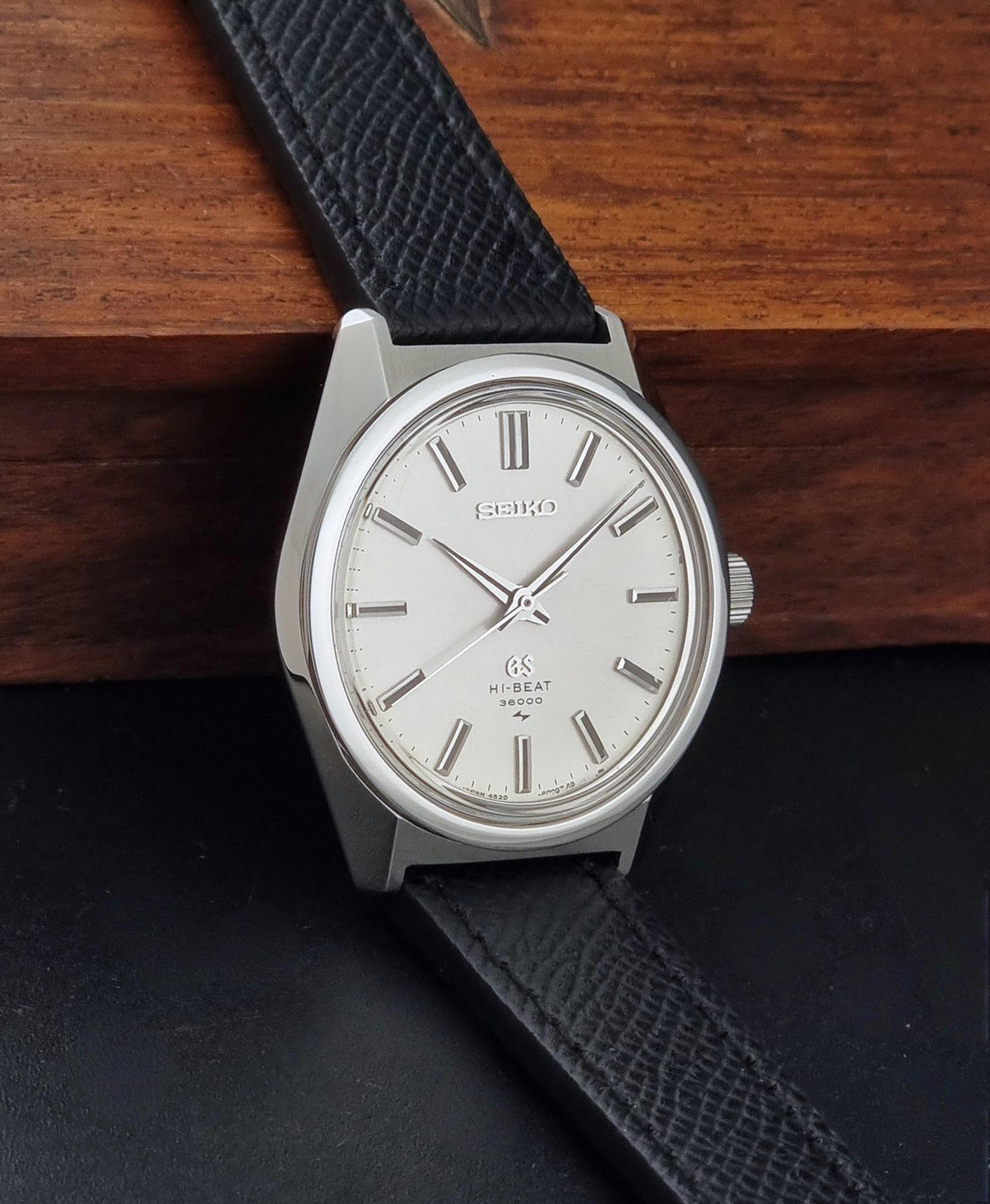 A vintage Grand Seiko watch 4520-8000 with a stainless steel case and black leather strap on a dark background resting on a wooden box